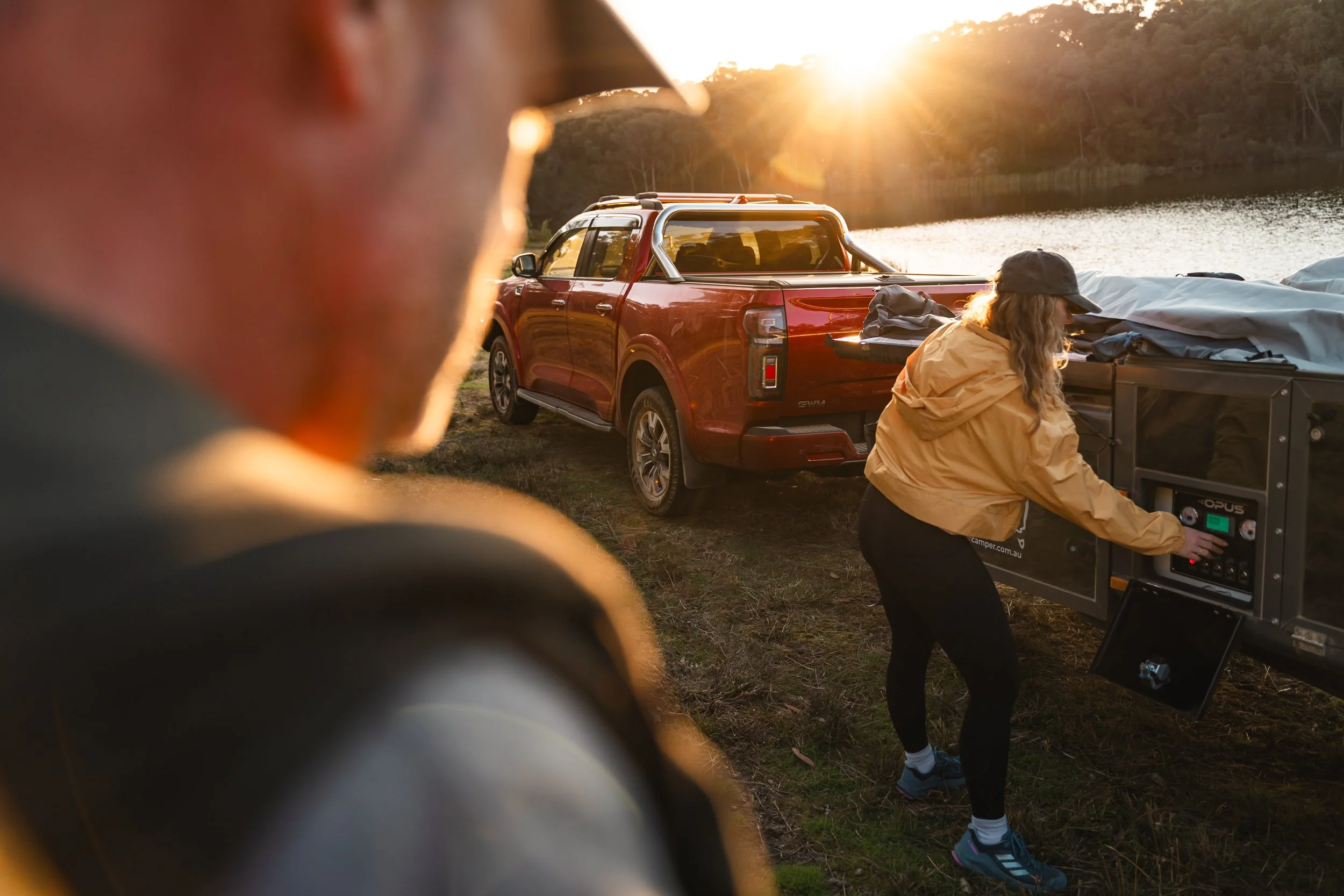 Person in the foreground with blurred face, two women near a red truck by a lakeside at sunset, one operating a generator, camping gear on the truck bed.