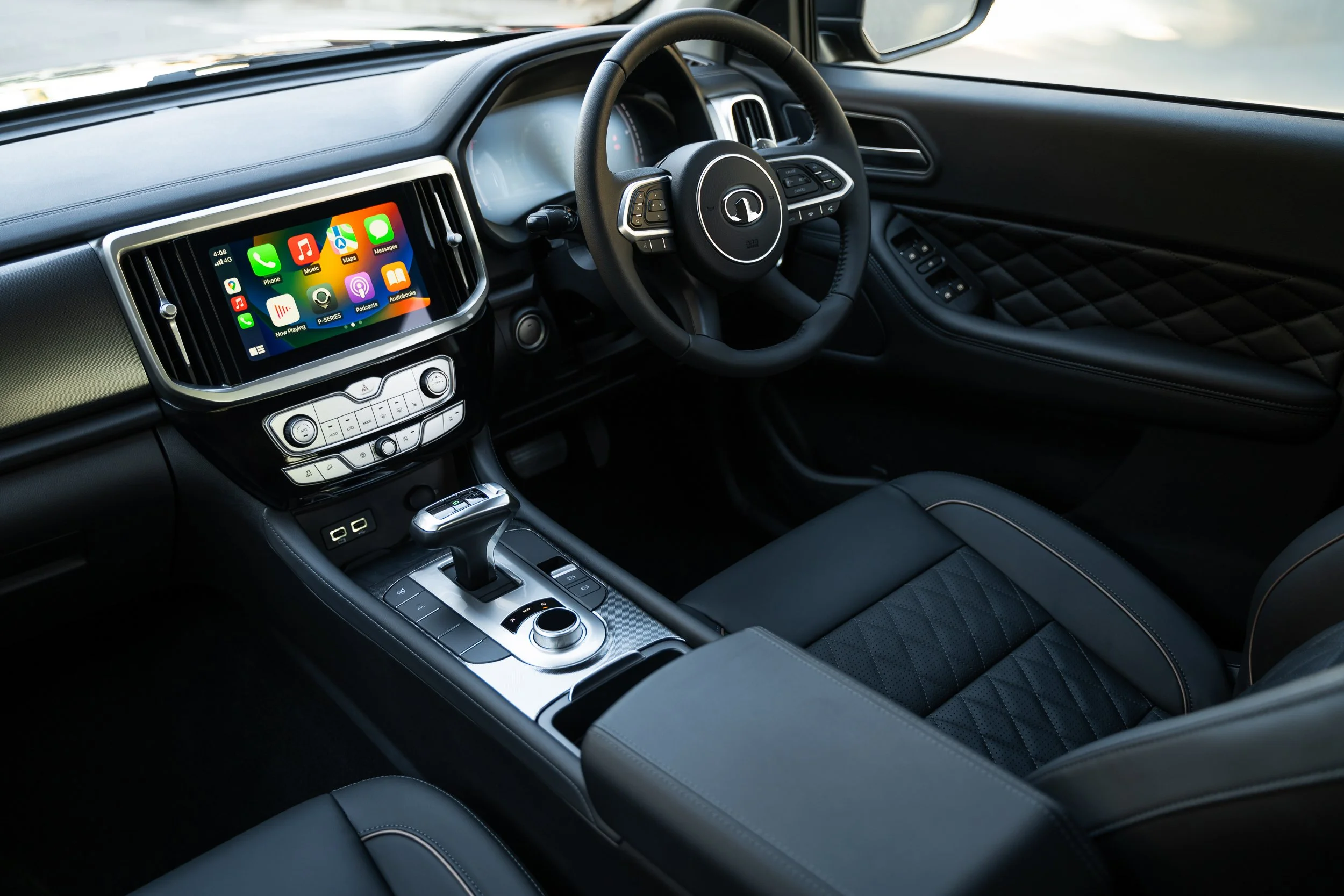 Interior view of a modern car dashboard featuring a digital touchscreen display showing various app icons, a steering wheel with control buttons, and climate control settings. The seats are black with quilted stitching.