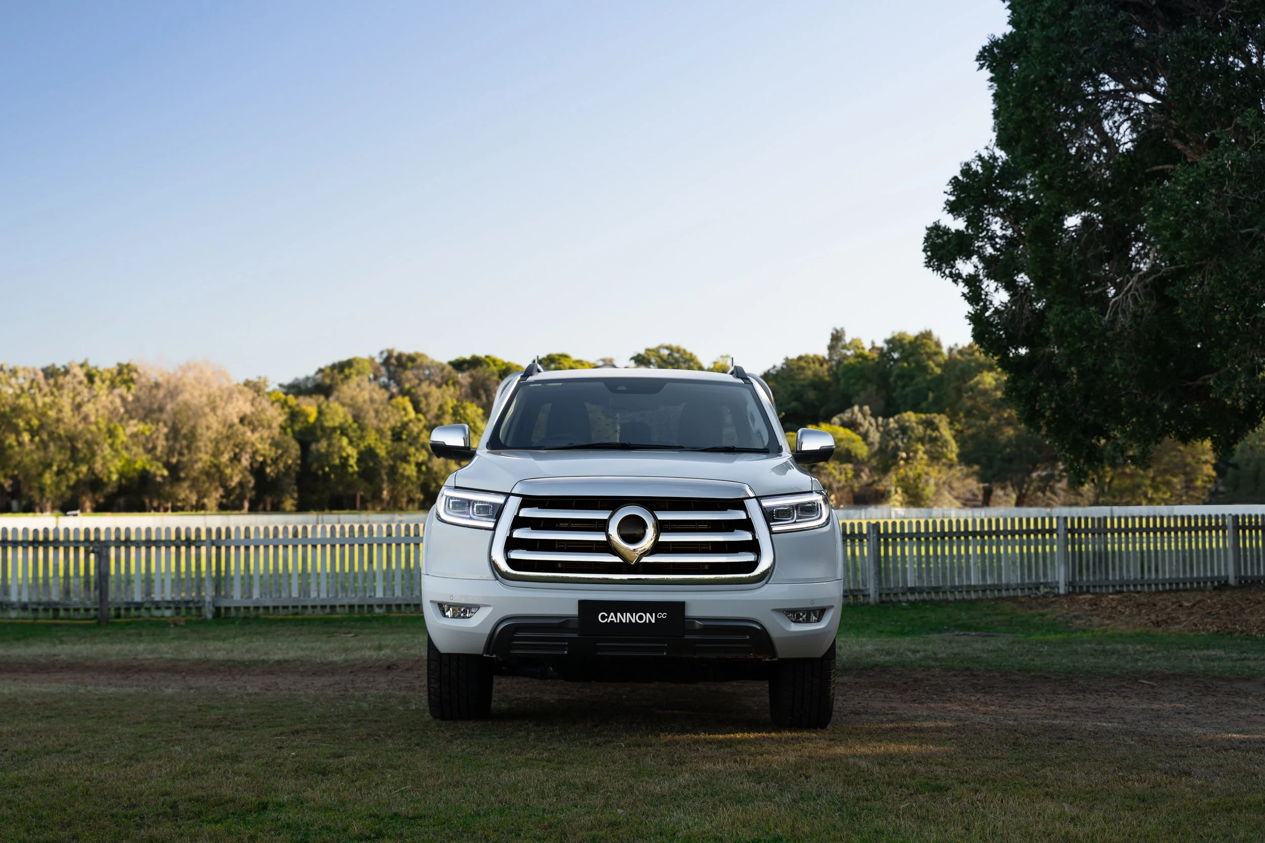 White SUV parked in front of a white picket fence of a iconic Australian cricket oval with trees in the background under a clear sky.