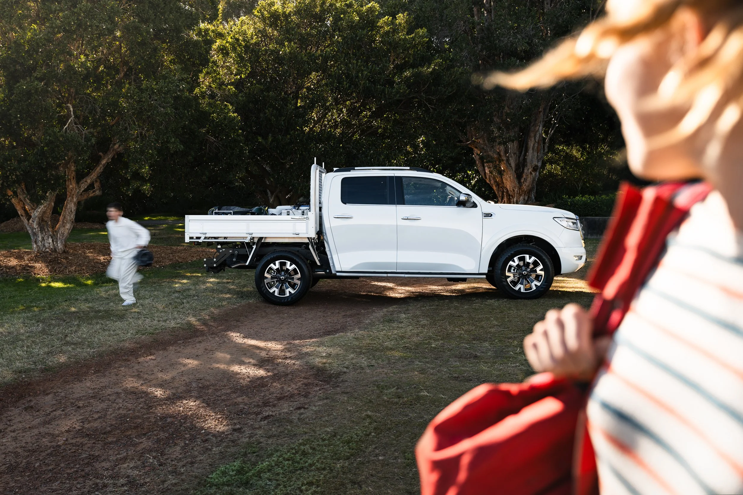 A white pickup truck parked on grass near trees, with a person blurred walking in the background and a child in the foreground. The child is wearing a striped shirt and appears to be holding or examining something.