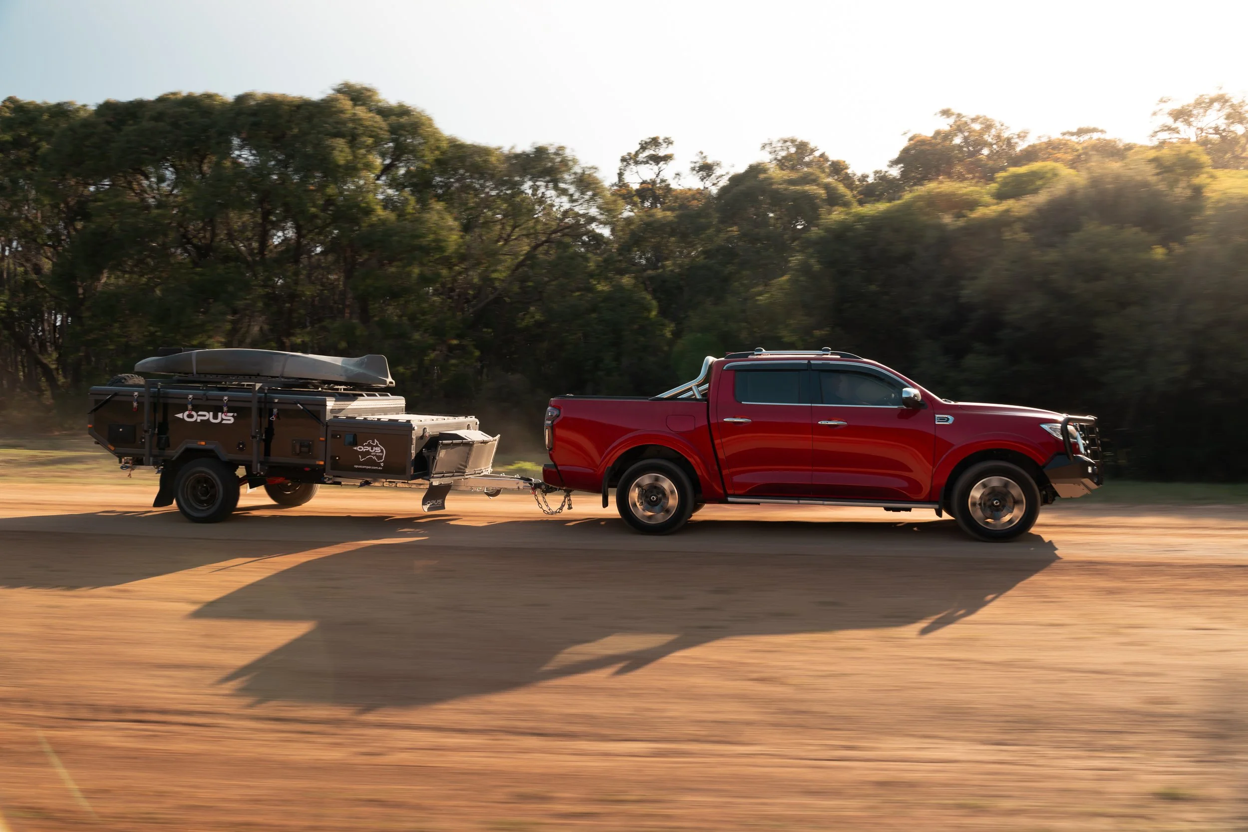 A red pickup truck towing a black trailer carrying a canoe on a dirt road surrounded by trees.