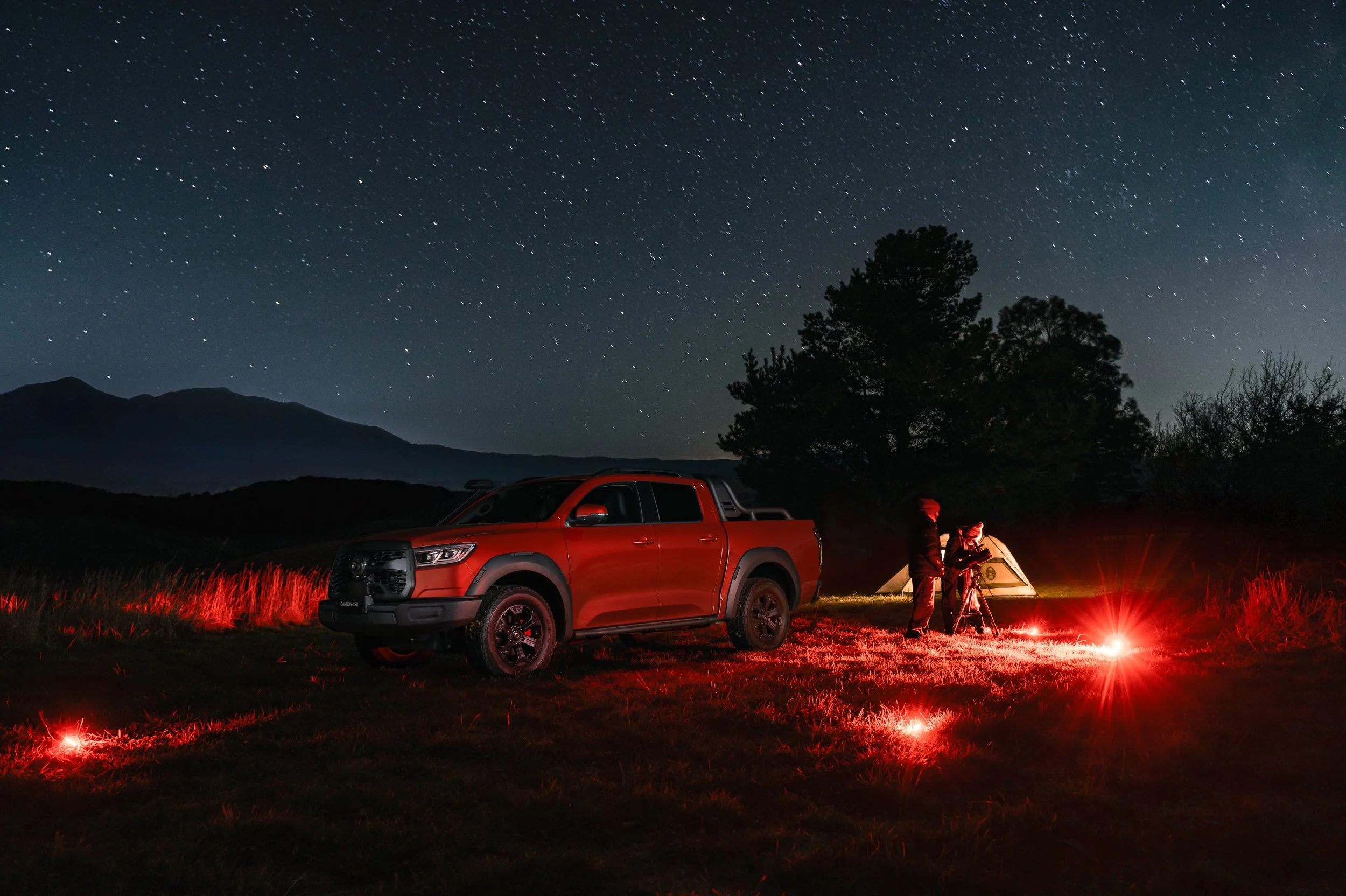A nighttime outdoor camping scene with a red pickup truck parked on grass, a tent, and two people using a telescope. The sky is filled with stars and other celestial objects, with mountain silhouettes in the background and a tree near the tent.