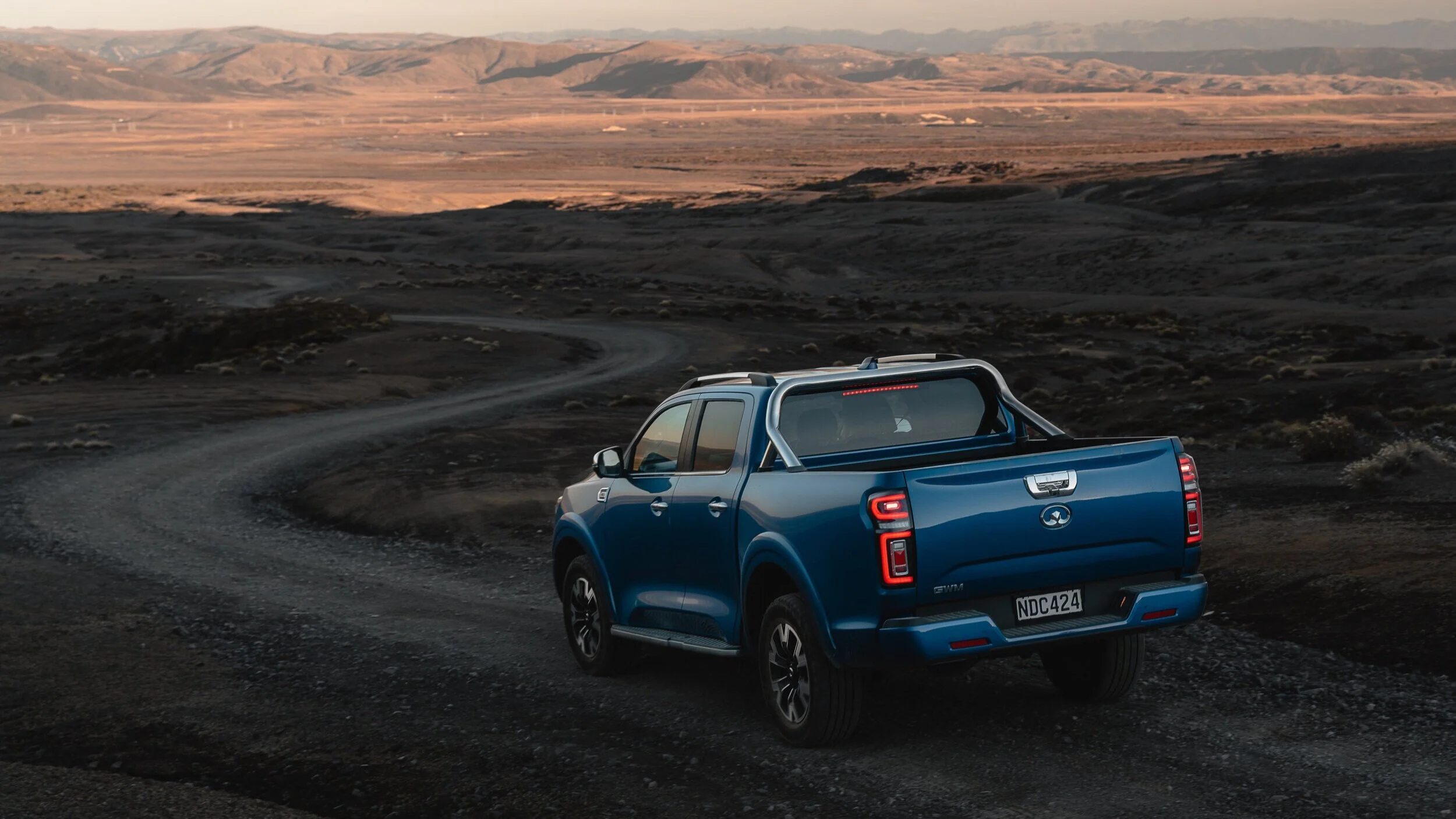 Blue pickup truck driving on a winding dirt road in a rural, mountainous landscape at sunset.