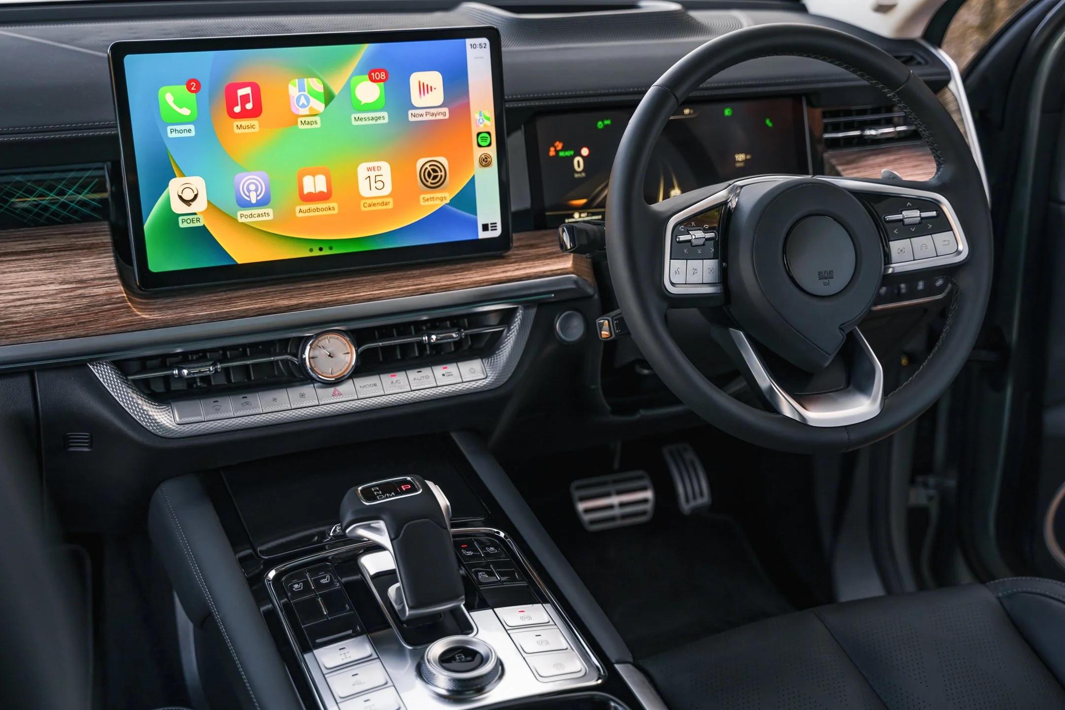 Inside view of a modern car dashboard featuring a large touchscreen display showing app icons for Phone, Music, Maps, Messages, Now Playing, Podcasts, Audiobooks, Calendar, and Settings. The steering wheel has various control buttons, and the center 
