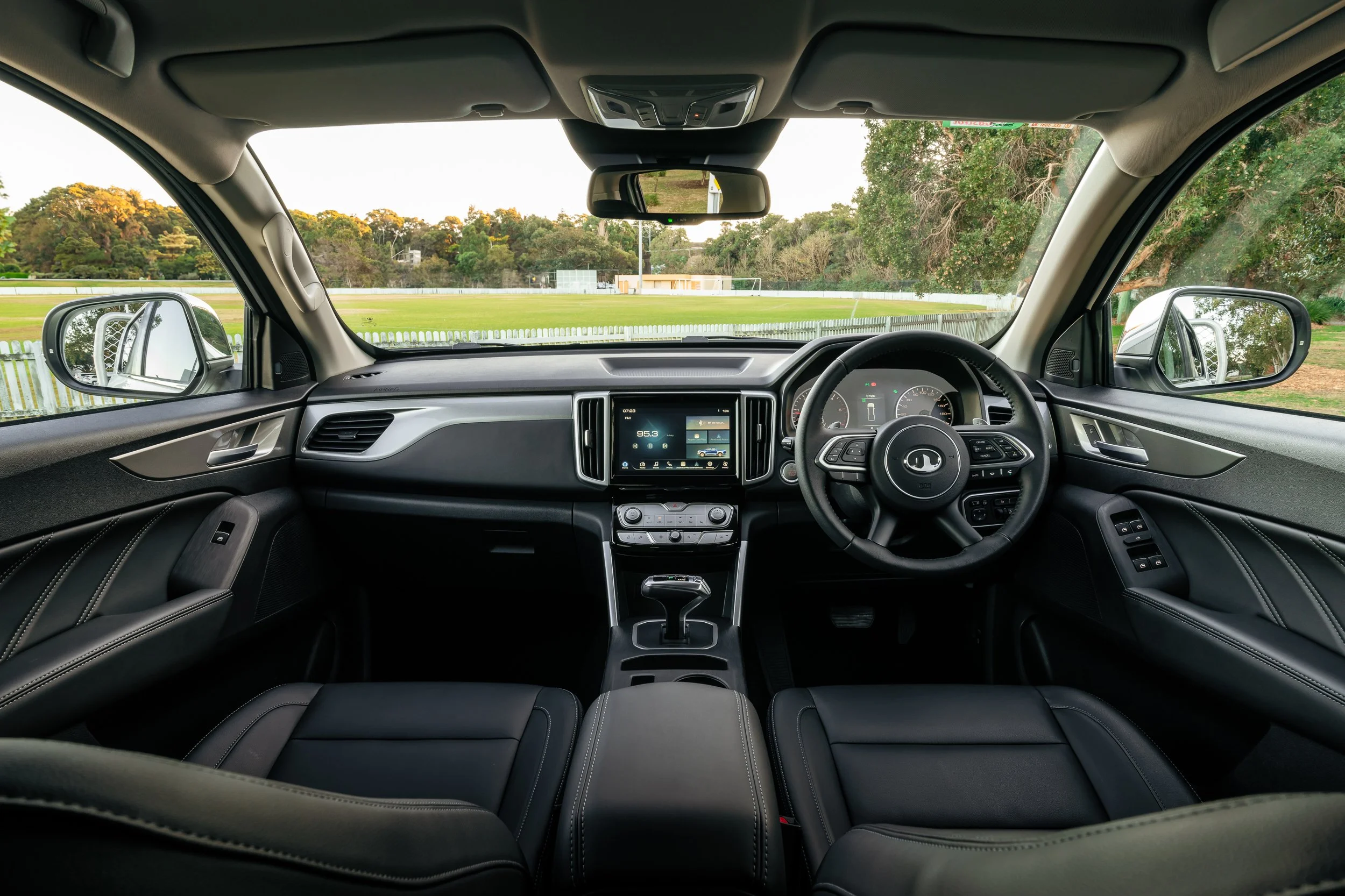 Inside view of a car looking through the front windshield, showing a green field with trees and a fence, with side mirrors reflecting the surroundings.
