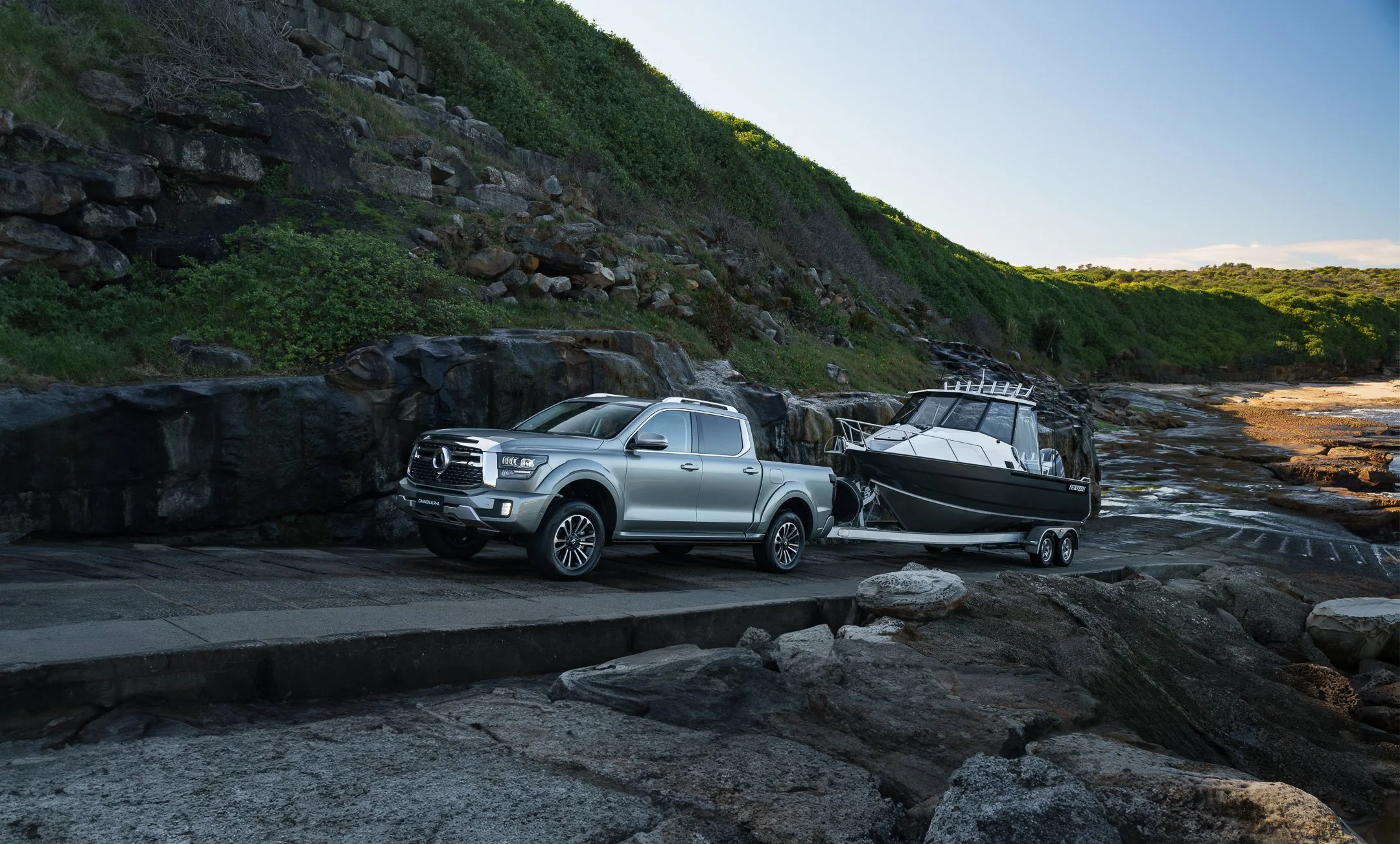 A silver pickup truck towing a boat on a trailer parked next to a rocky shoreline with a hillside covered in greenery, during daylight.