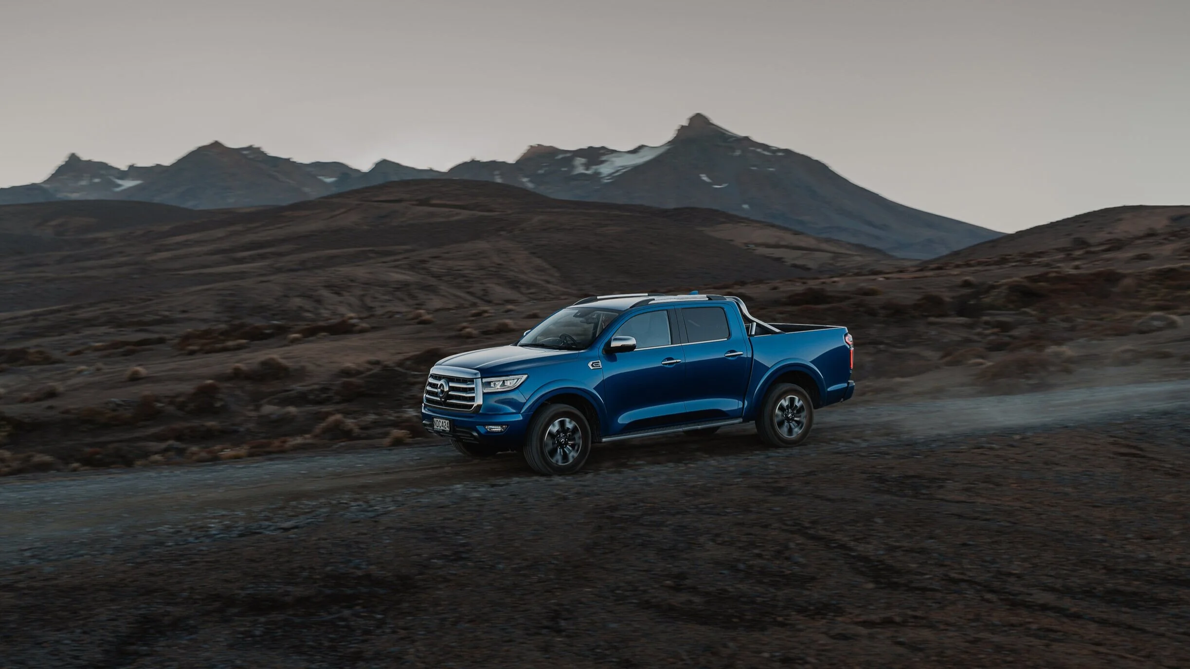 A blue pickup truck driving on a dirt road in a barren mountainous landscape with snow-capped peaks in the background during dusk or dawn.