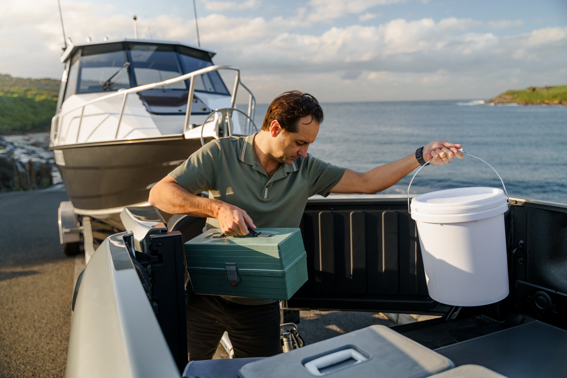 A man on a boat dock working with fishing equipment, with a boat, water, and cloudy sky in the background.