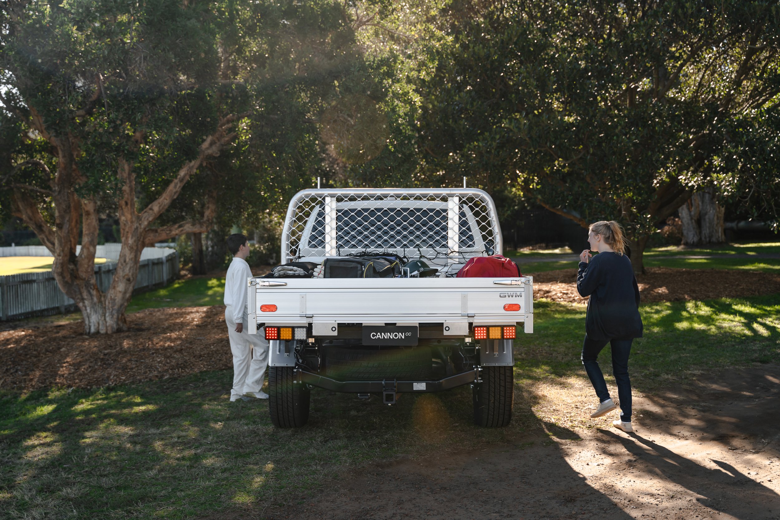 A man and a woman stand outside near the back of a white pickup truck filled with various bags and gear, under trees with sunlight filtering through
