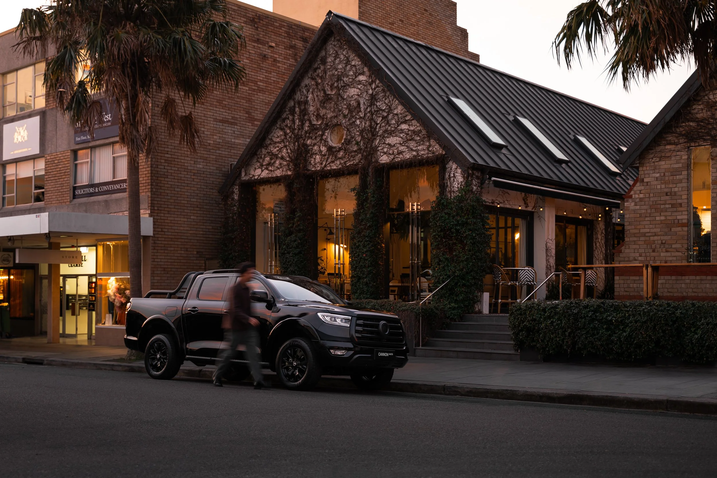 A city street scene at dusk with a black pickup truck parked in front of a building with large glass doors. A person is walking past the truck. The building has brick walls, large windows, and a shadow of a vine on the front. There are palm trees on 