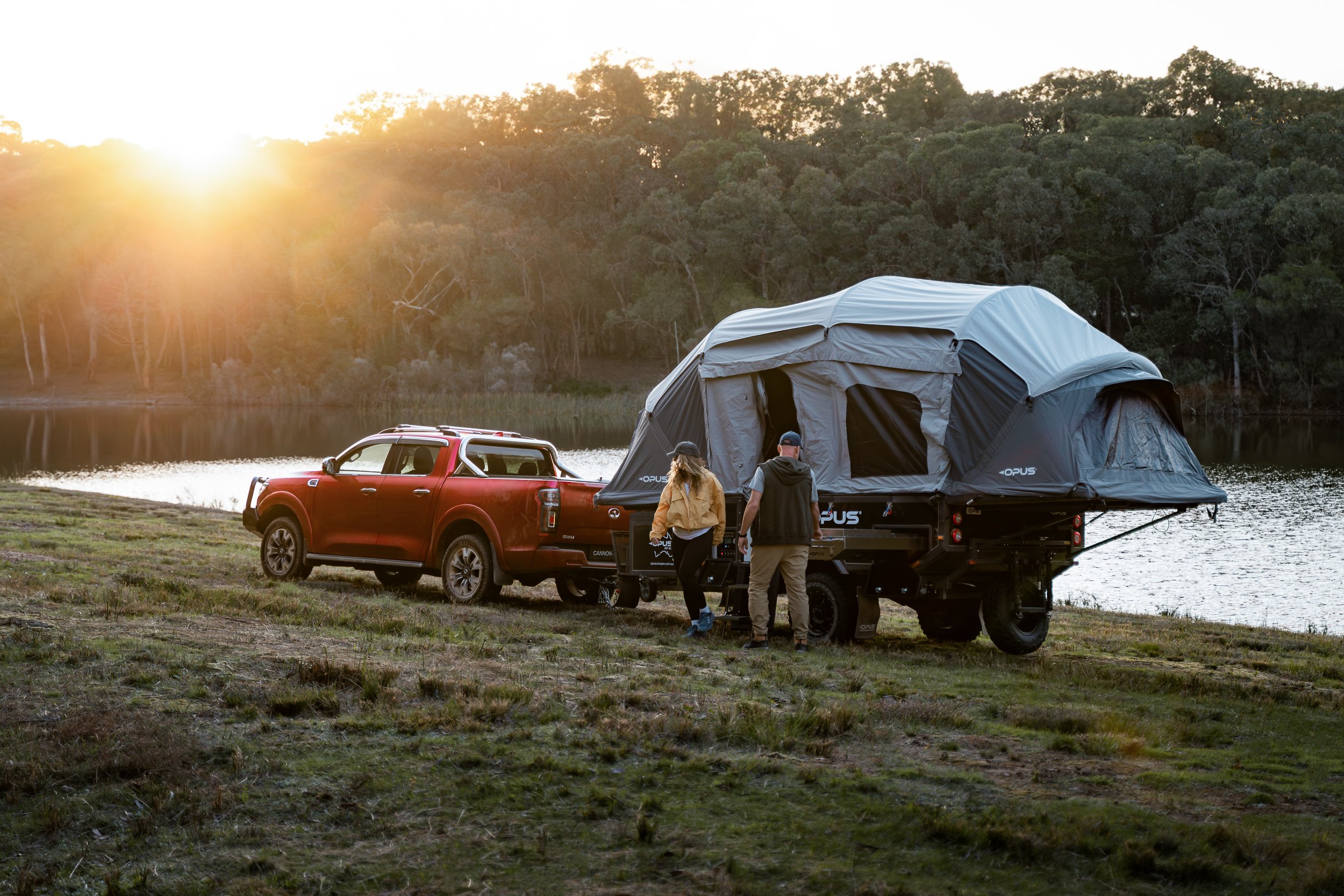 A red pickup truck parked beside a lake at sunset with a large camping tent mounted on a trailer behind. Three people are near the trailer, one is adjusting the tent, and the others are standing nearby, all outdoors surrounded by trees.