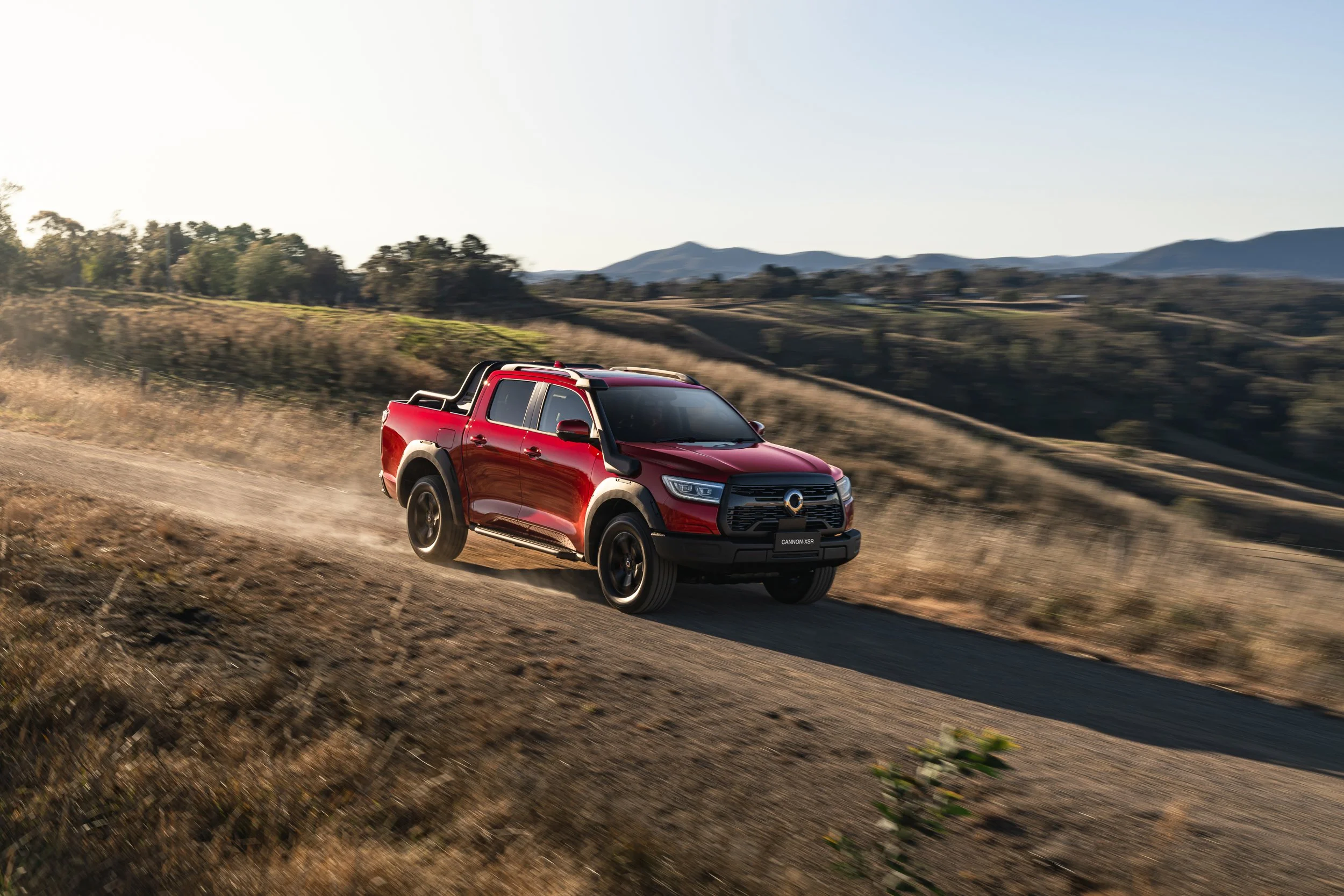 Red pickup truck driving through a rural landscape with rolling hills and mountains in the background during late afternoon or early evening.