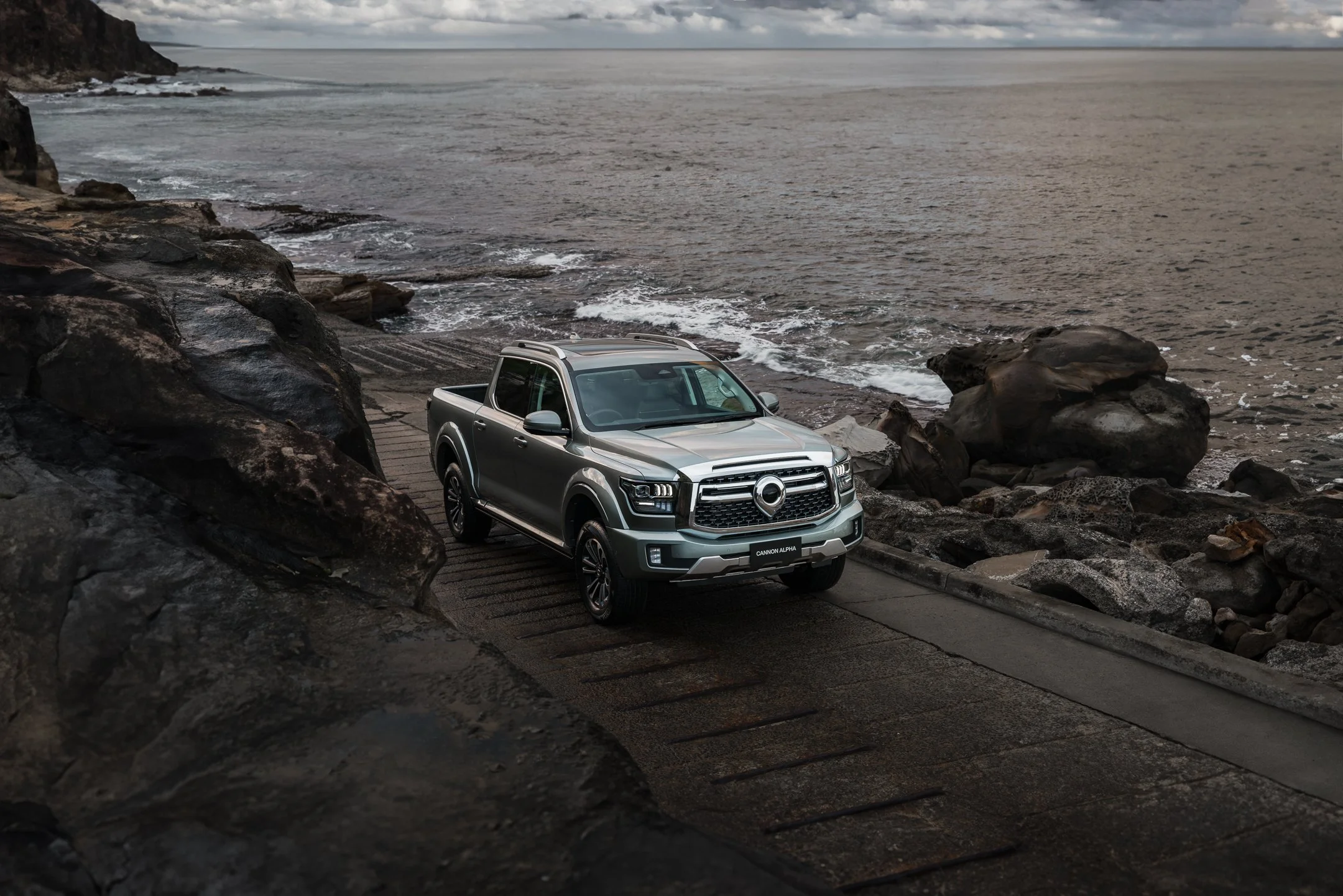 A silver pickup truck parked on a paved area near a rocky shoreline with ocean water and cloudy sky in the background.
