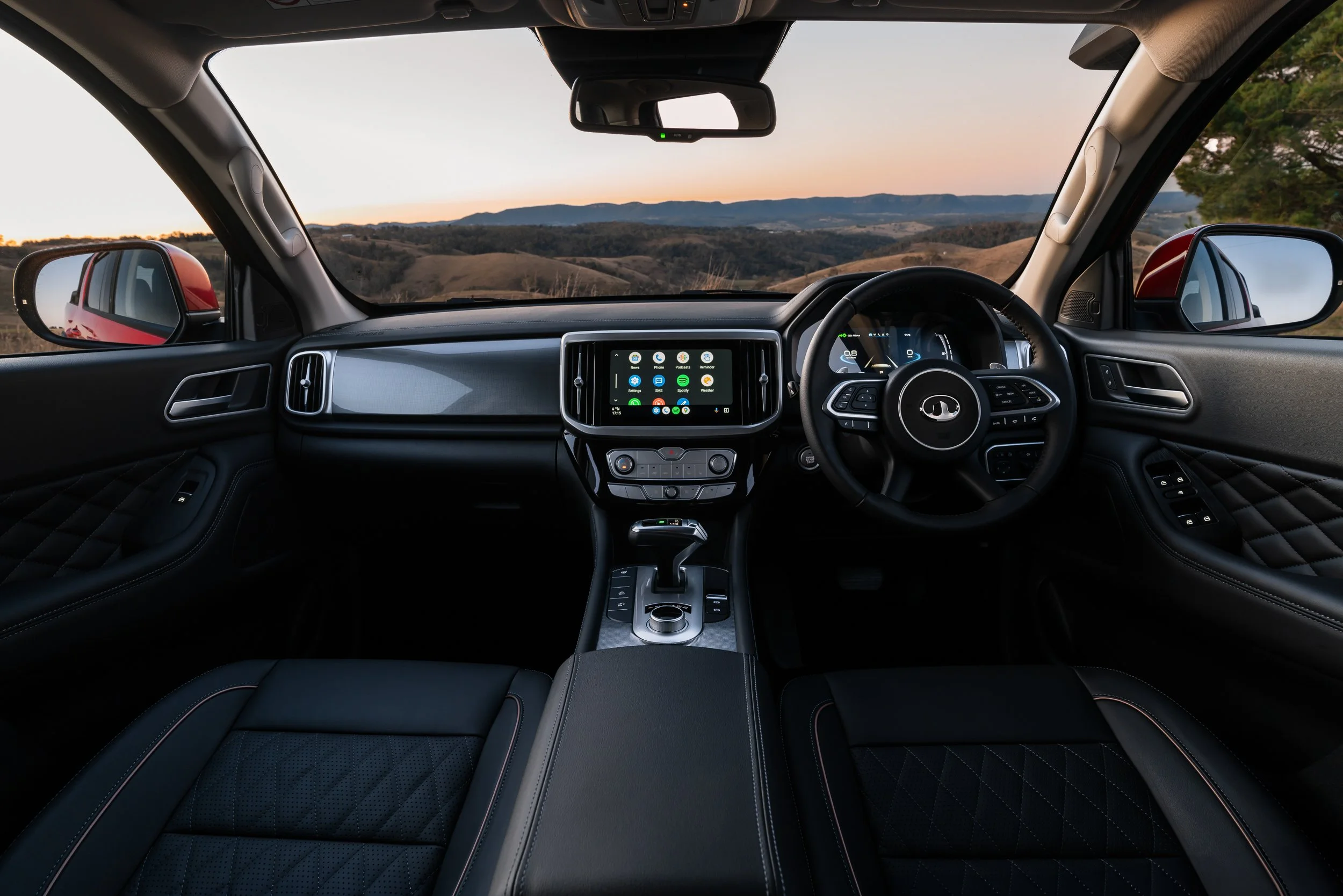 The interior of a modern car showing the dashboard, steering wheel, and infotainment screen, with a scenic view of rolling hills and a sunset through the windshield.