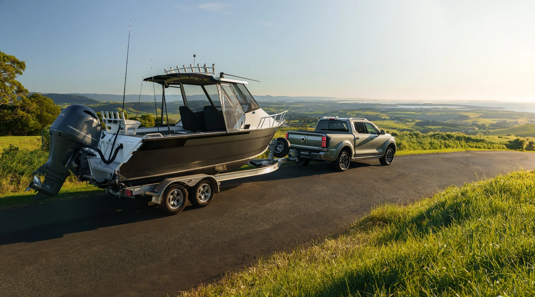 A boat on a trailer attached to a pickup truck, parked on a scenic overlook with green rolling hills and water in the distance under a clear sky.