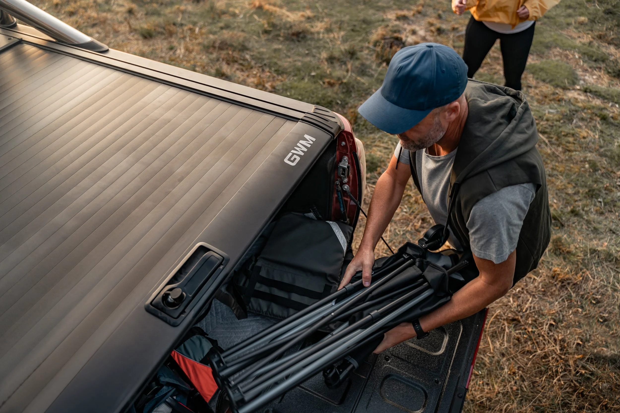 A man wearing a blue cap and gray hoodie is packing equipment into the trunk of a vehicle, with a woman in a tan jacket and black leggings standing outside nearby, on a grassy, leaf-strewn area.