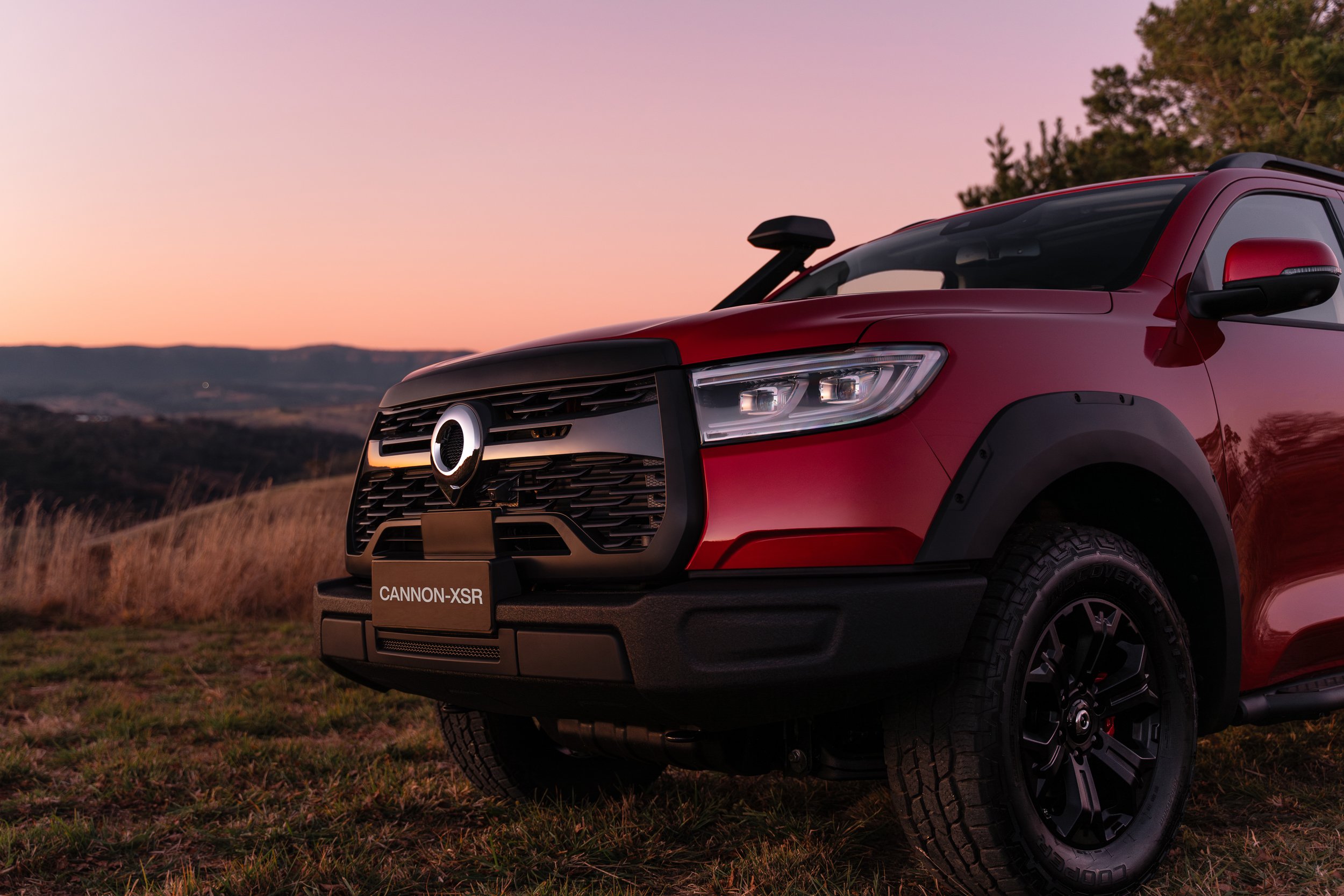 Red off-road vehicle parked on grassy field at sunset with rolling hills and trees in the background.