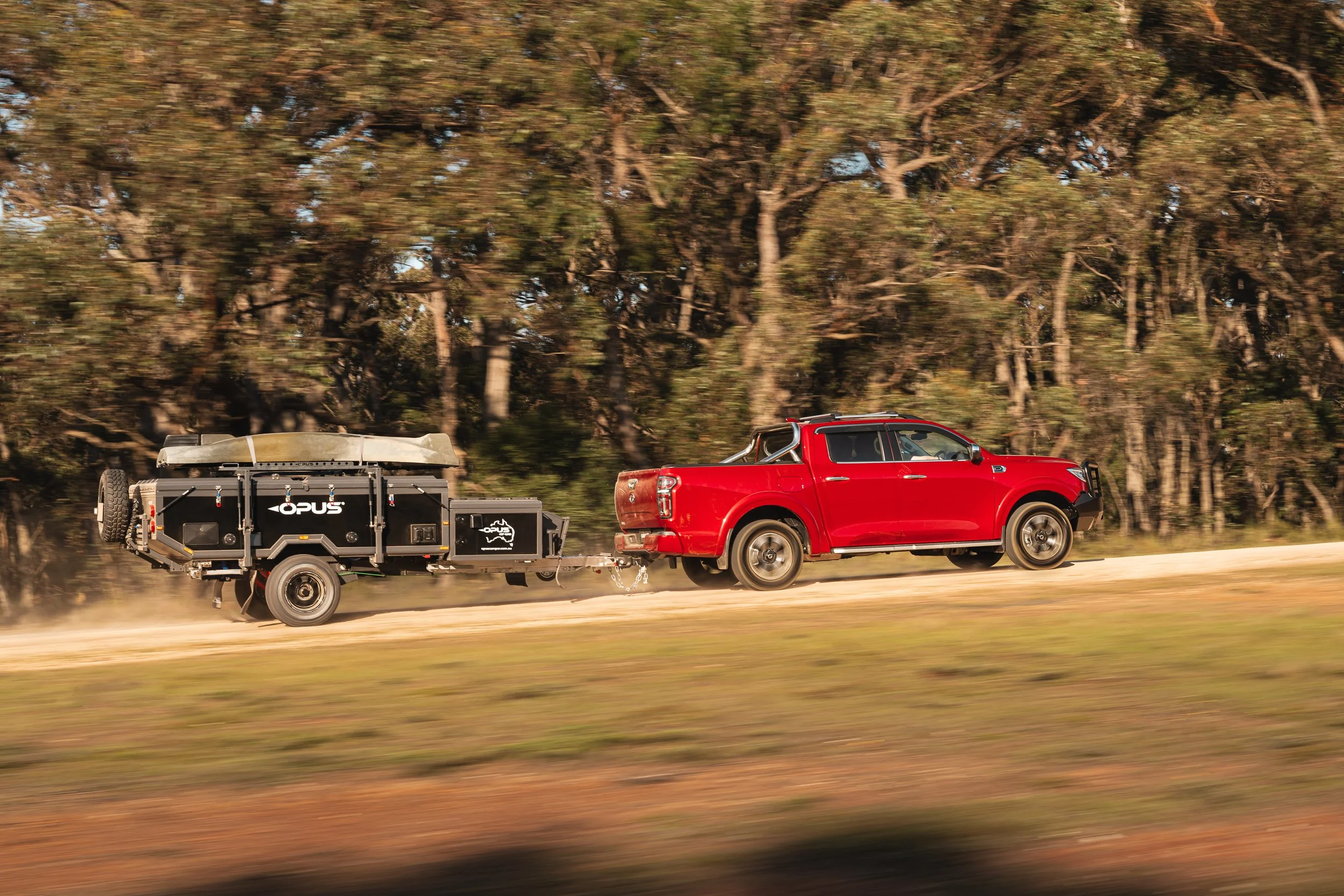 Red pickup truck towing a trailer through a blurred landscape of trees and dusty road.