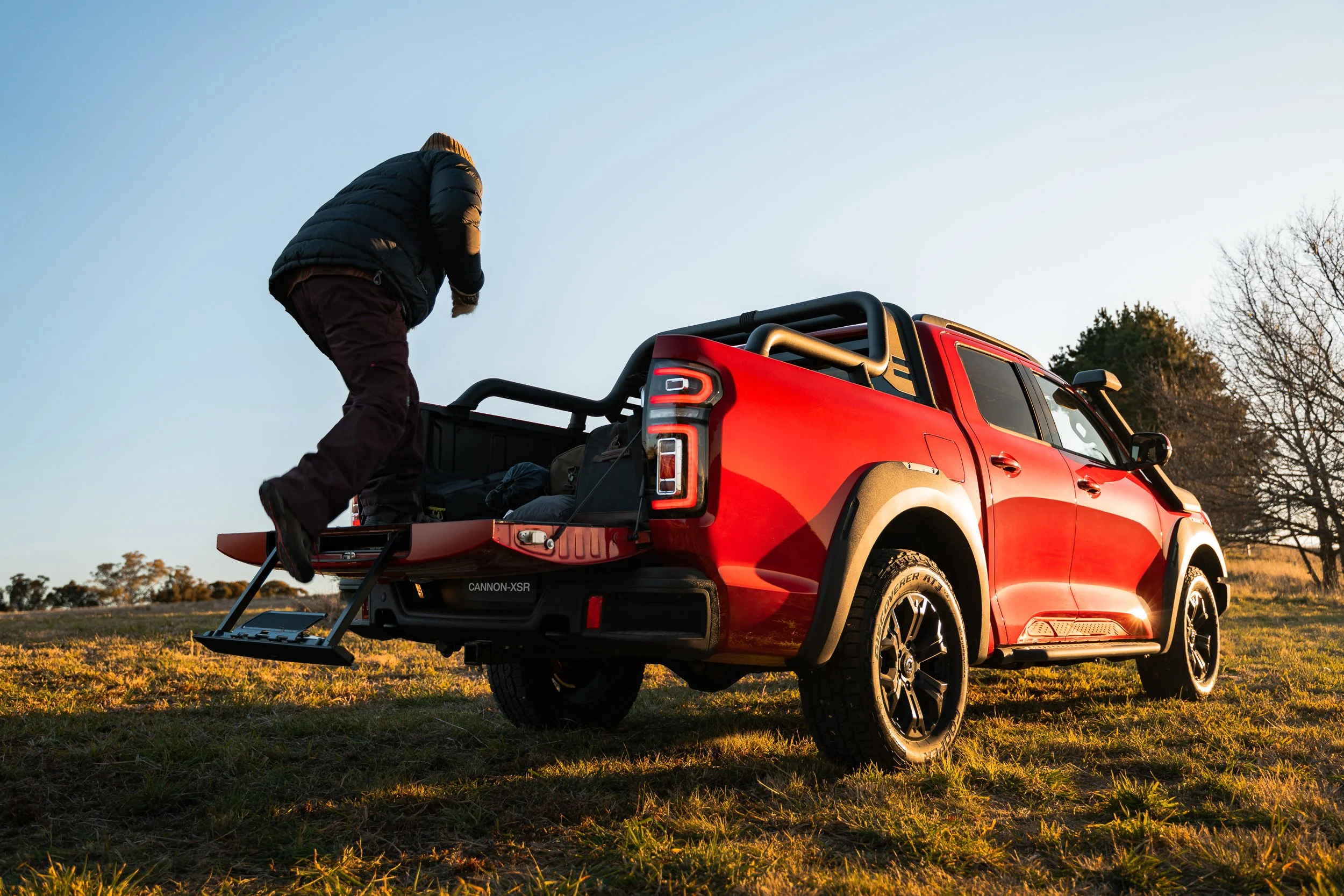 Person loading gear into the back of a red pickup truck parked on a grassy field during sunset with trees in the background.