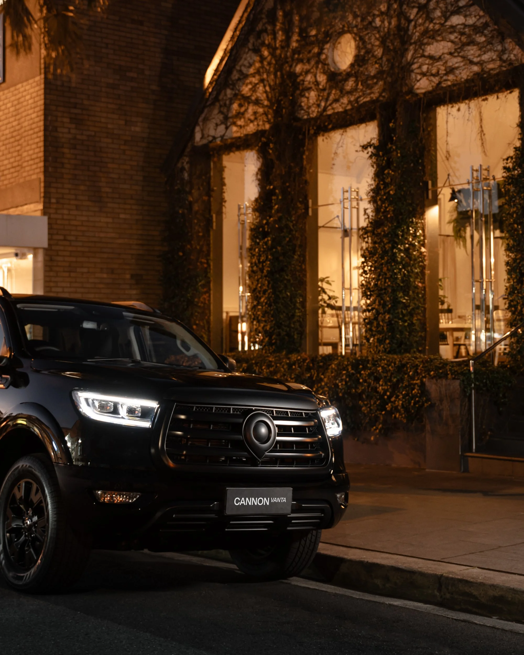 Close-up of a black car with a Cannon Vanta license plate parked on a street at night. In the background, there is a building with ivy-covered walls, illuminated by warm lighting.