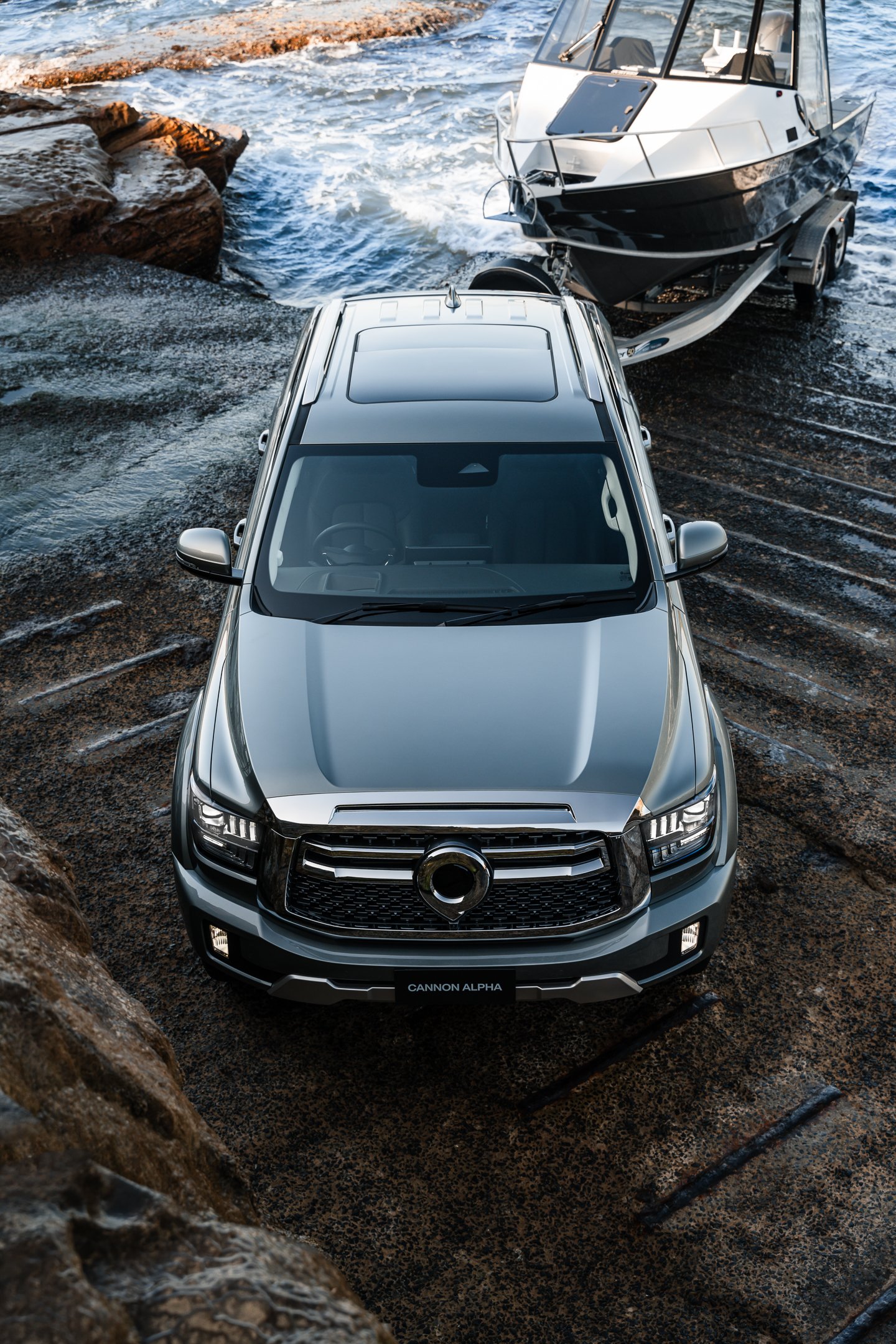 A silver SUV with a black grille and a "Cannon Alpha" license plate is parked on rocky terrain near water, with a boat on a trailer in the background.