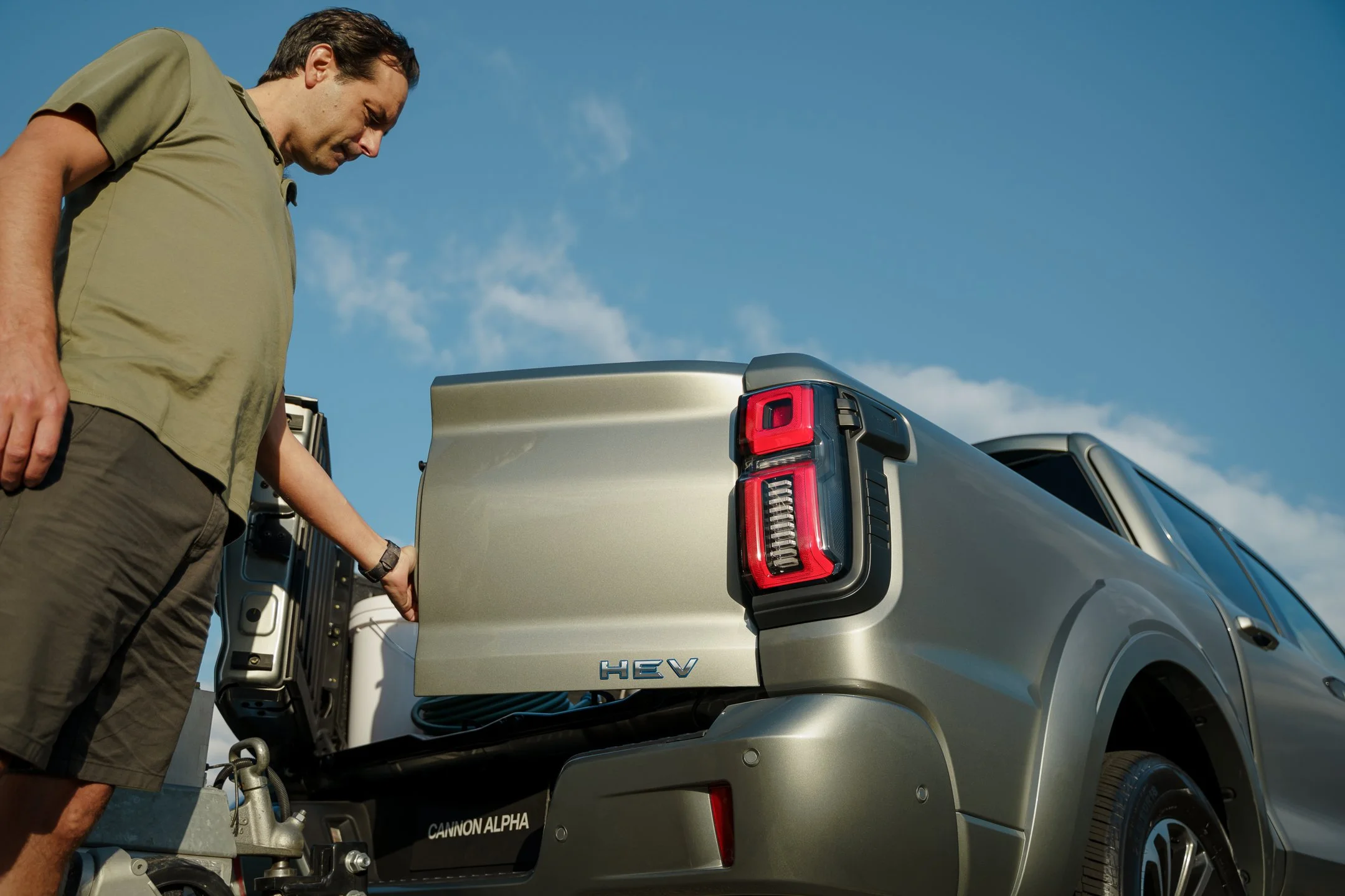 A man in a tan shirt and shorts standing next to a silver electric vehicle with its trunk open, revealing an equipment compartment, under a clear blue sky.