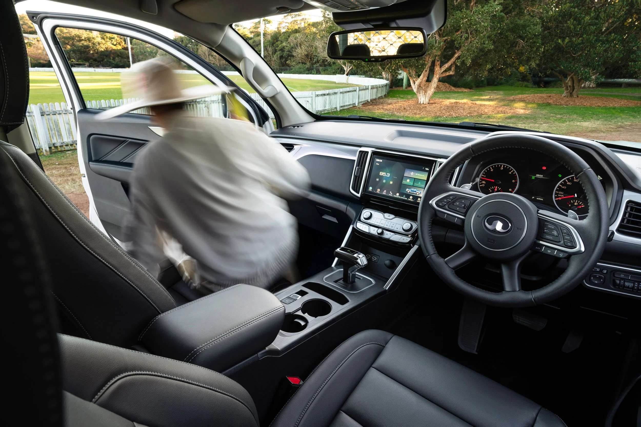 View from inside a vehicle showing the dashboard, steering wheel, and front seats. The passenger, a young cricket player, rushes out of the car excited to play cricket at the oval
