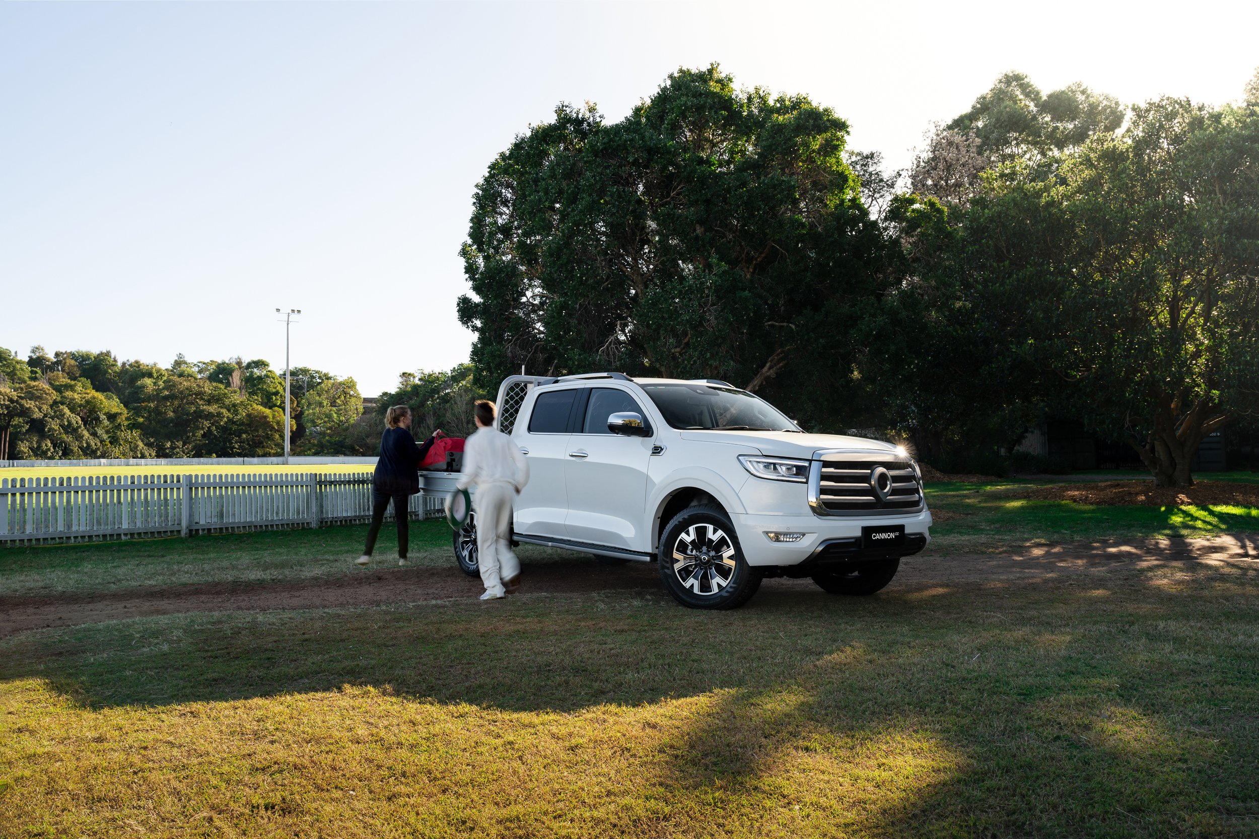 Two women are standing beside a white pickup truck with the name 'CANNON' on the front license plate. They are outdoors near a large green tree with bright sunlight shining through the leaves. One woman is wearing white and the other is dressed in da