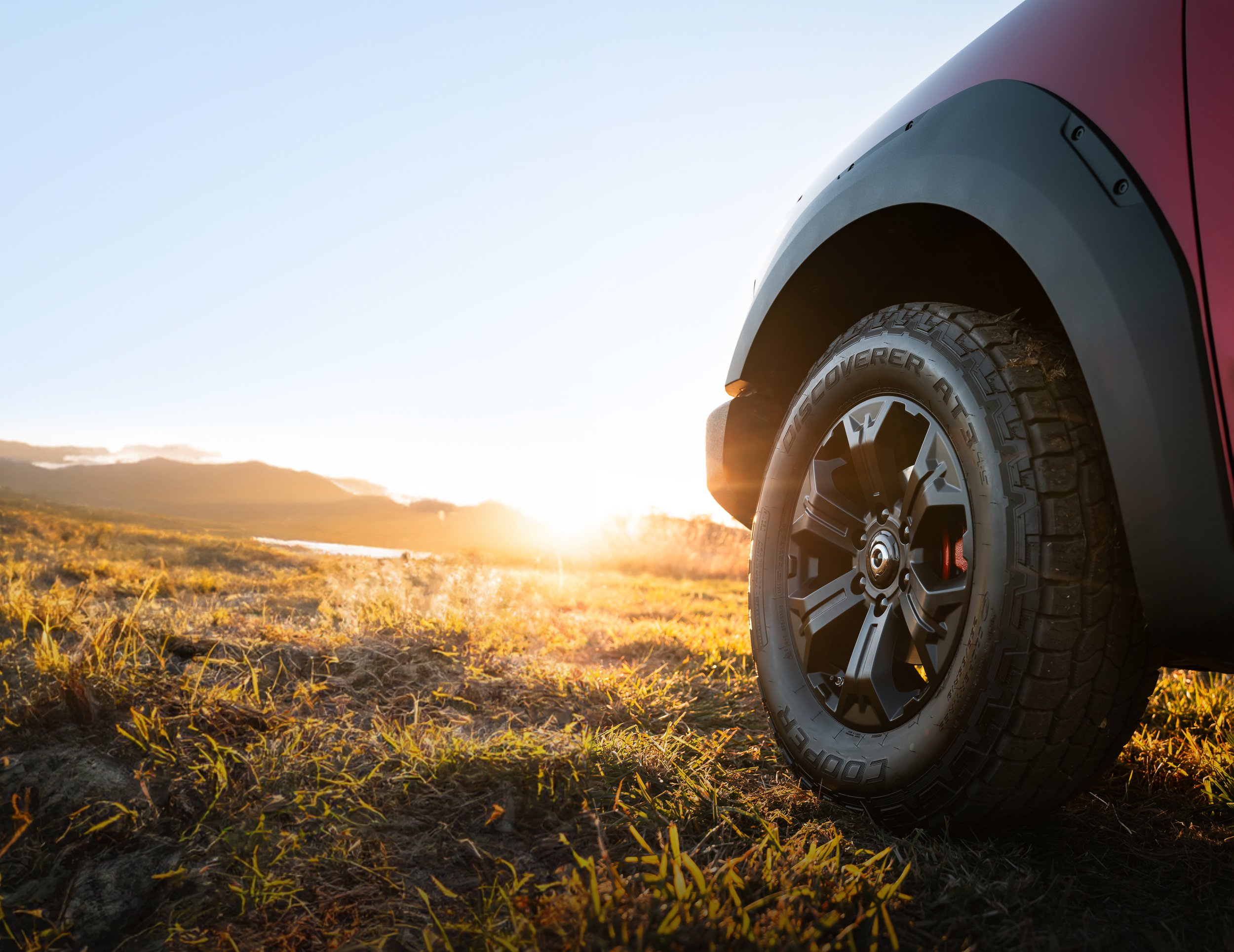 Close-up of a red off-road vehicle's tire and wheel, with a sunset over a grassy landscape in the background.