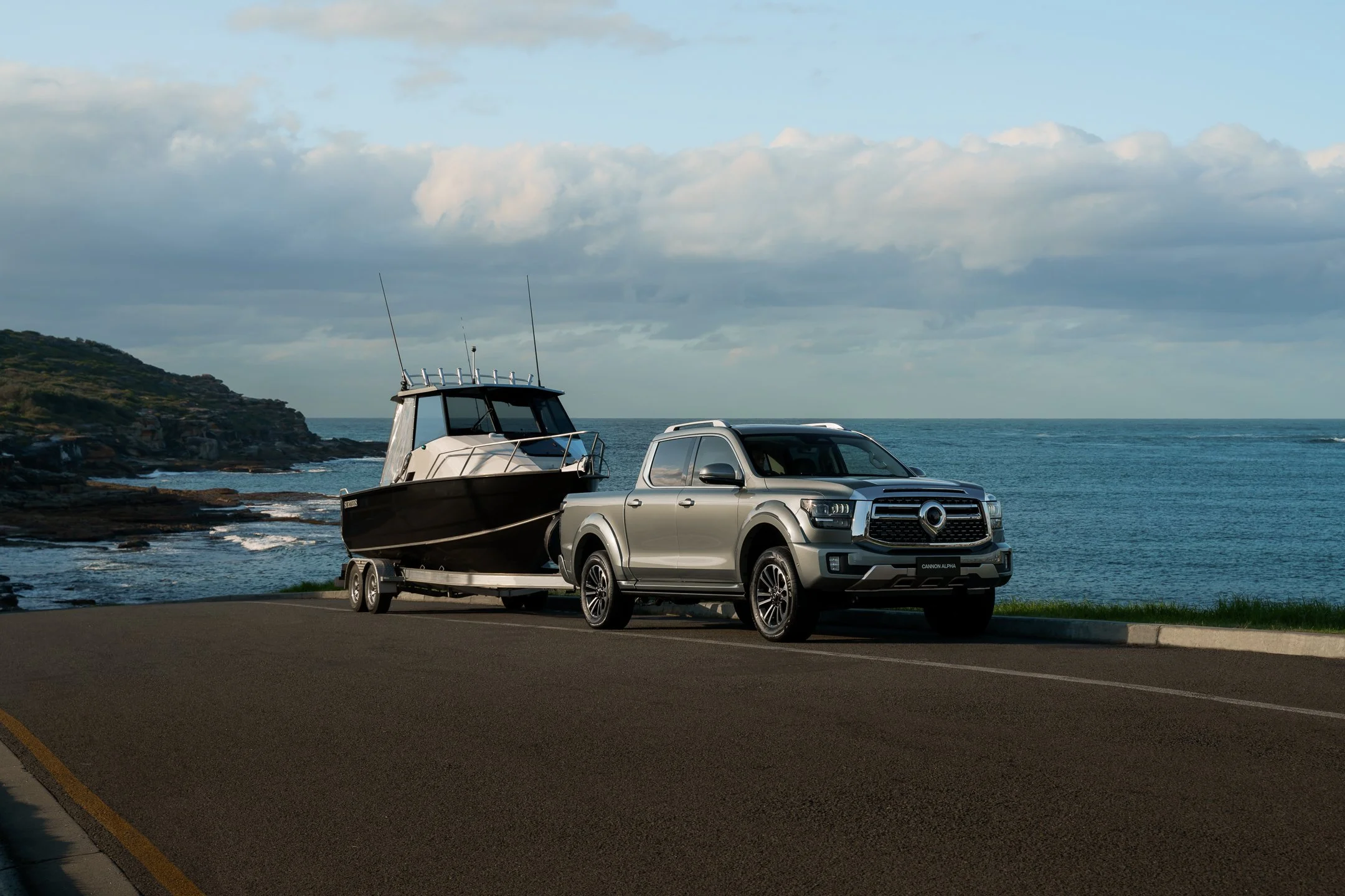 A silver pickup truck towing a boat on a trailer along a paved road near the ocean with rocky shores and a hill in the background under a cloudy sky.