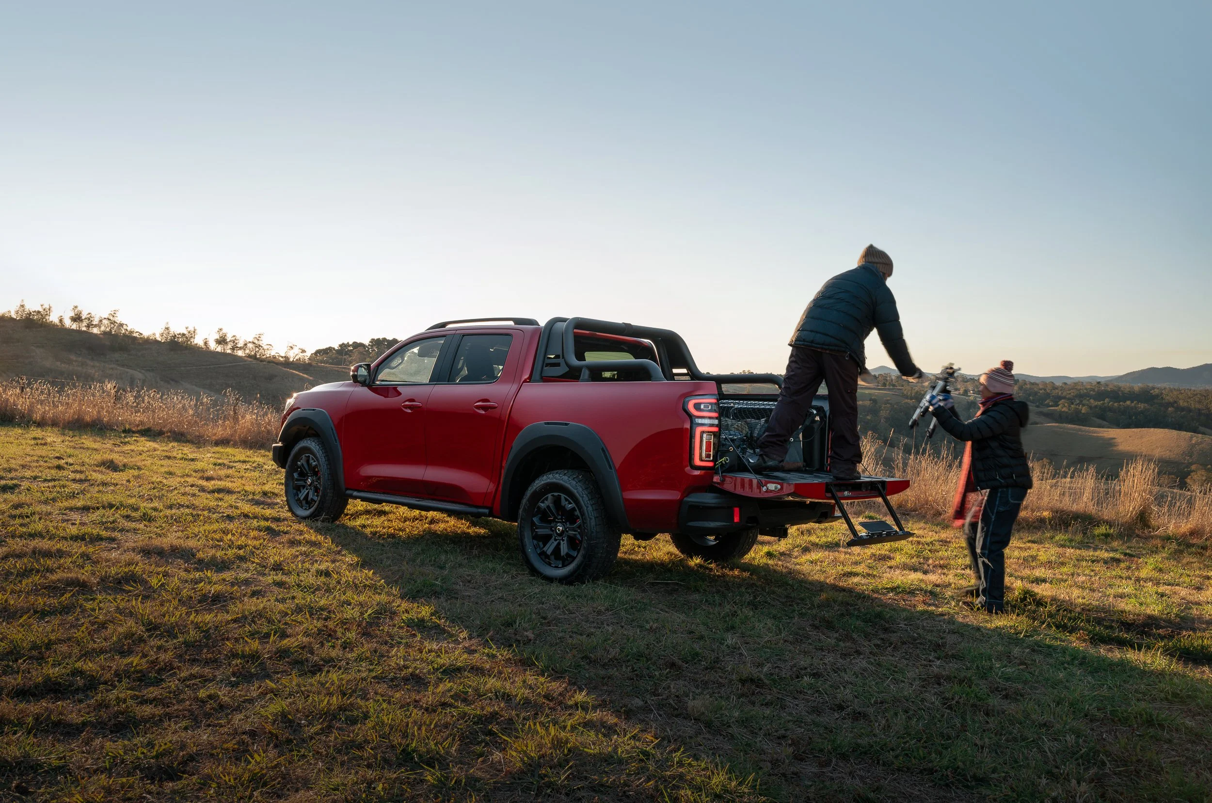 A red pickup truck parked in a grassy field with hills in the background at sunset. Two children are by the truck, one standing on the tailgate, the other holding a skateboard, both dressed warmly with hats and jackets.
