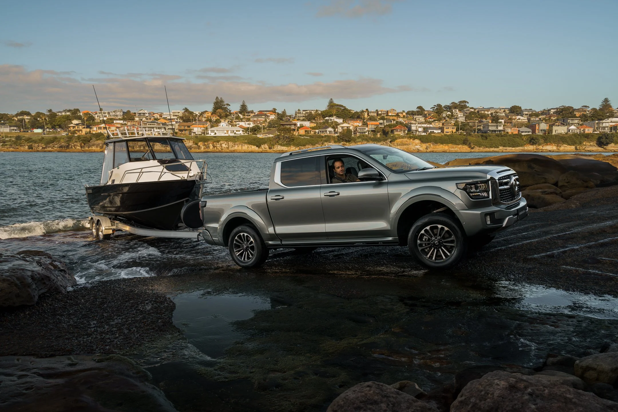 A silver pickup truck on a rocky beach carrying a boat on a trailer, with a coastal neighborhood in the background under a partly cloudy sky.