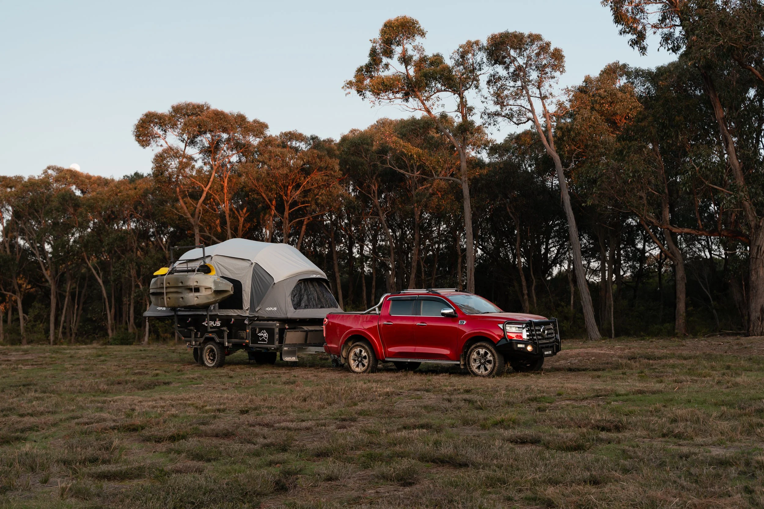 Red pickup truck parked on a grassy field, attached to a trailer carrying an outdoor tent and a kayak, with a dense forest of tall trees in the background during sunset.
