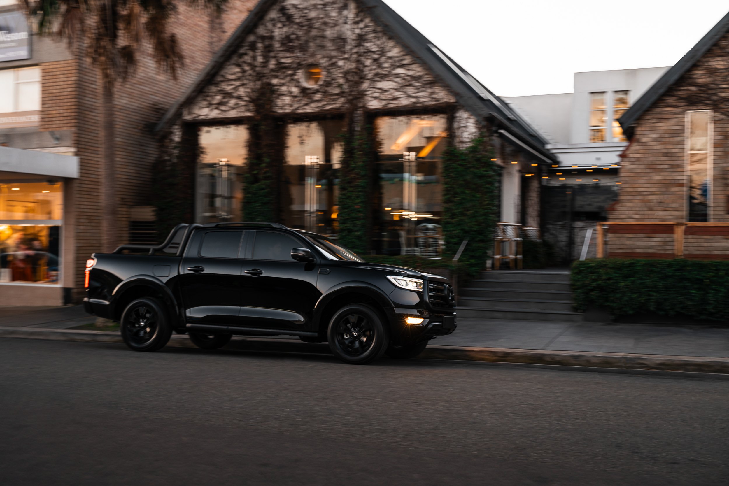 A black vehicle is driving on a city street in front of a modern building with large glass windows, brick walls, and outdoor string lights during evening or dusk.