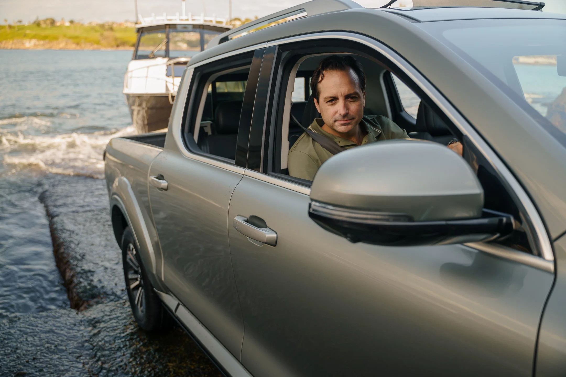 A man sits in the driver's seat of a silver pickup truck near a body of water with boats docked nearby. The man appears to be looking at the camera. The background includes a shoreline with grass and some buildings.