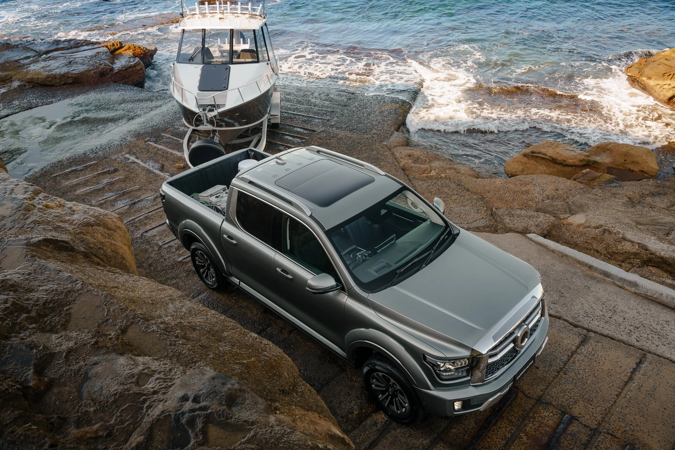 A silver pickup truck parked on a rocky shoreline with a boat on a trailer just above the waterline, close to the ocean with waves gently hitting the rocks.
