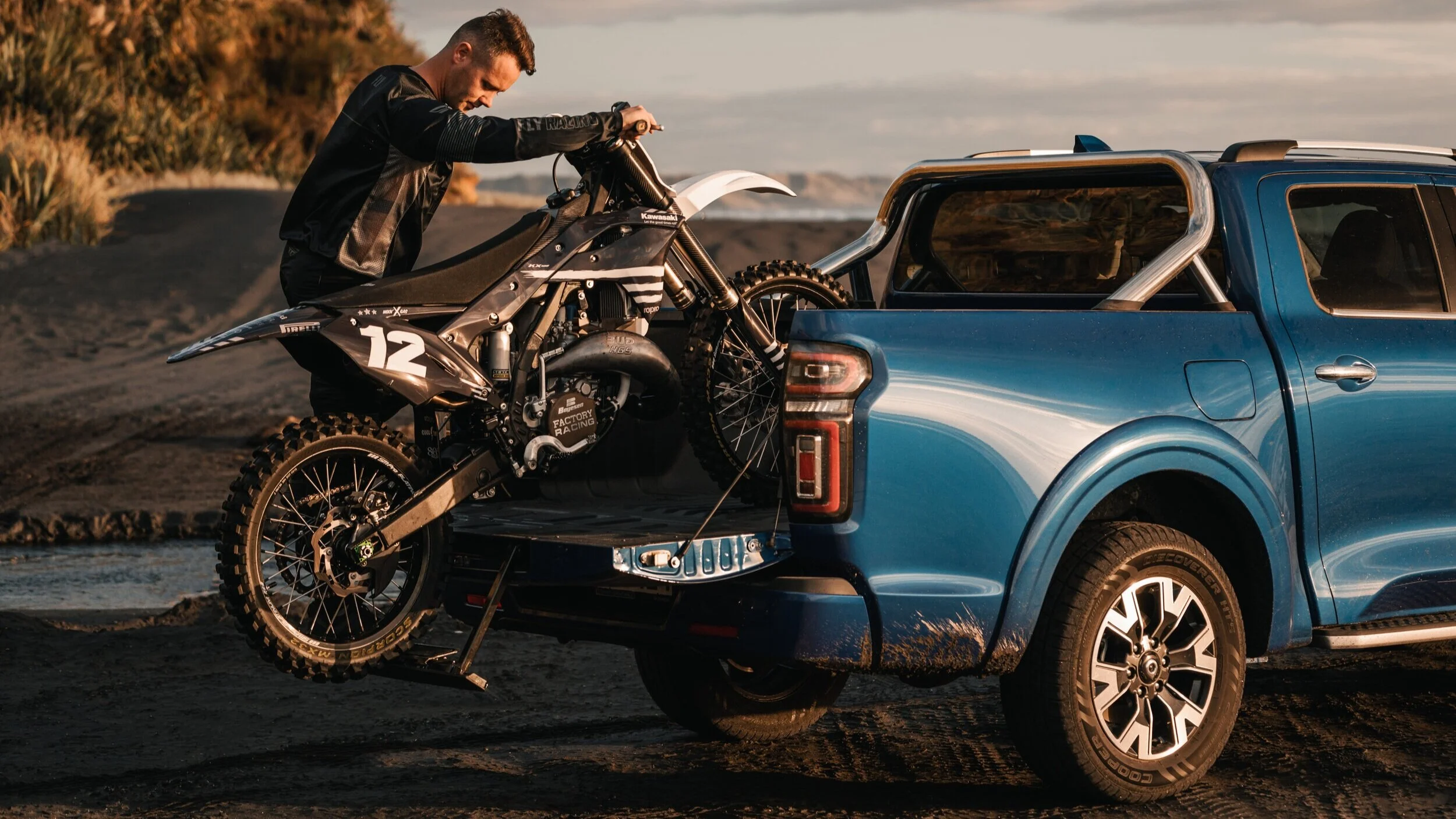 A man loading a dirt bike into the back of a blue pickup truck on a dirt road during sunset.