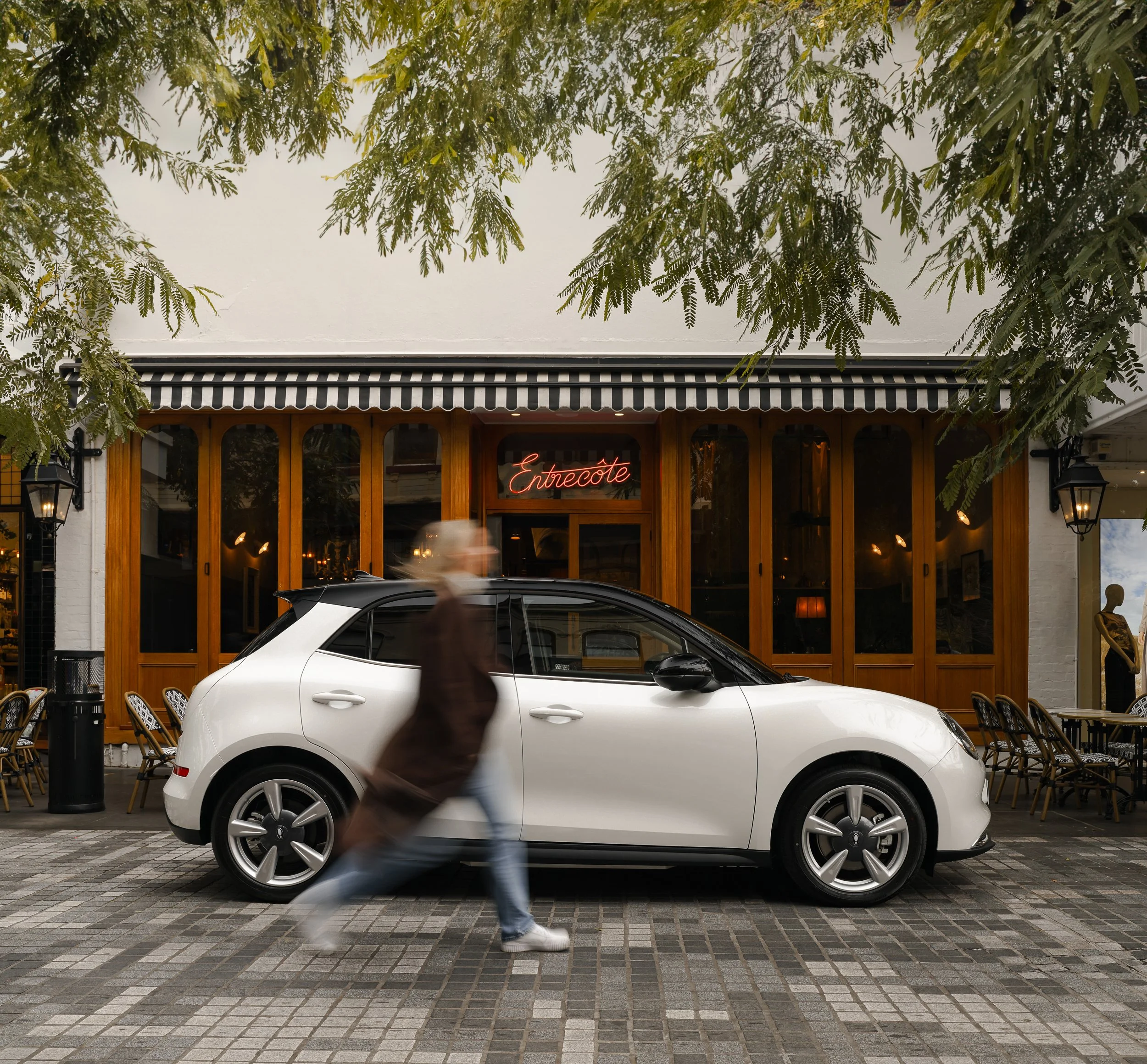 A white car driving down a city street with a person walking in front of it, slightly blurred.