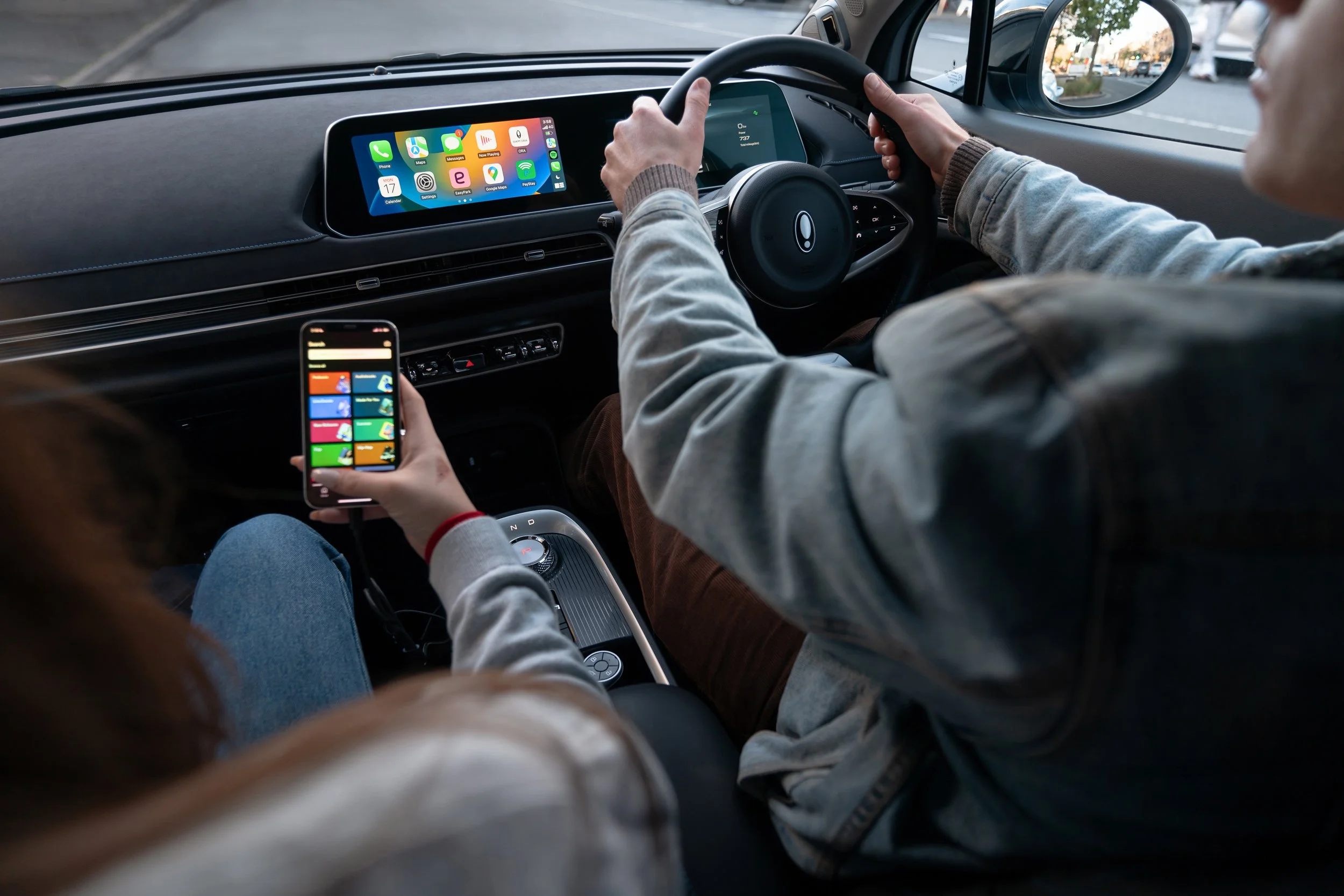 Interior view of a car with two passengers, one holding a smartphone with colorful app icons and the other driving, showing a digital dashboard and a city street outside.
