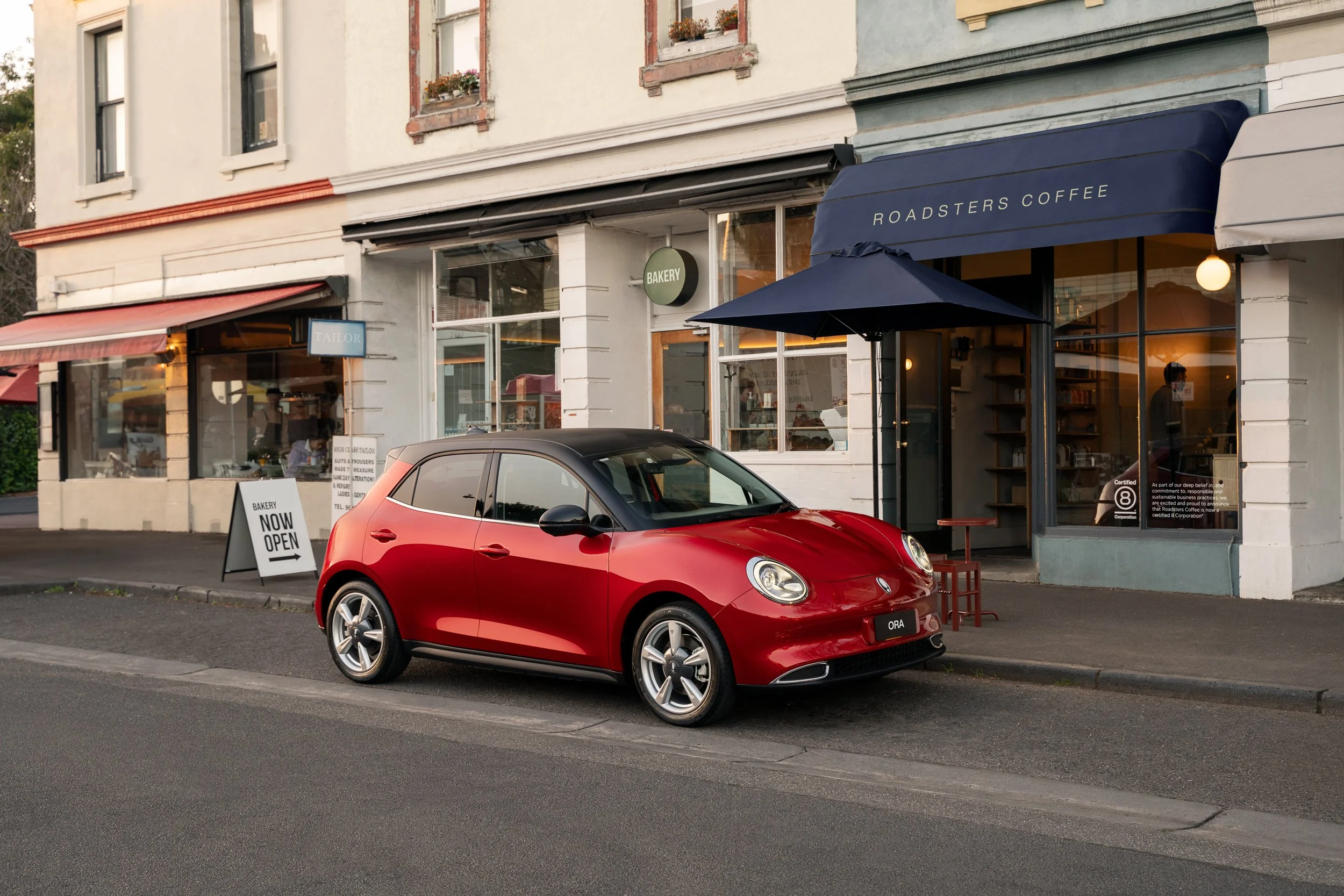 A red compact car parked in front of a storefront with large glass windows, signs for a bakery and tailoring shop, and a blue awning with the words 'Roadsters Coffee' on it. The storefront features a sidewalk sign that says 'Bakery Now Open.'