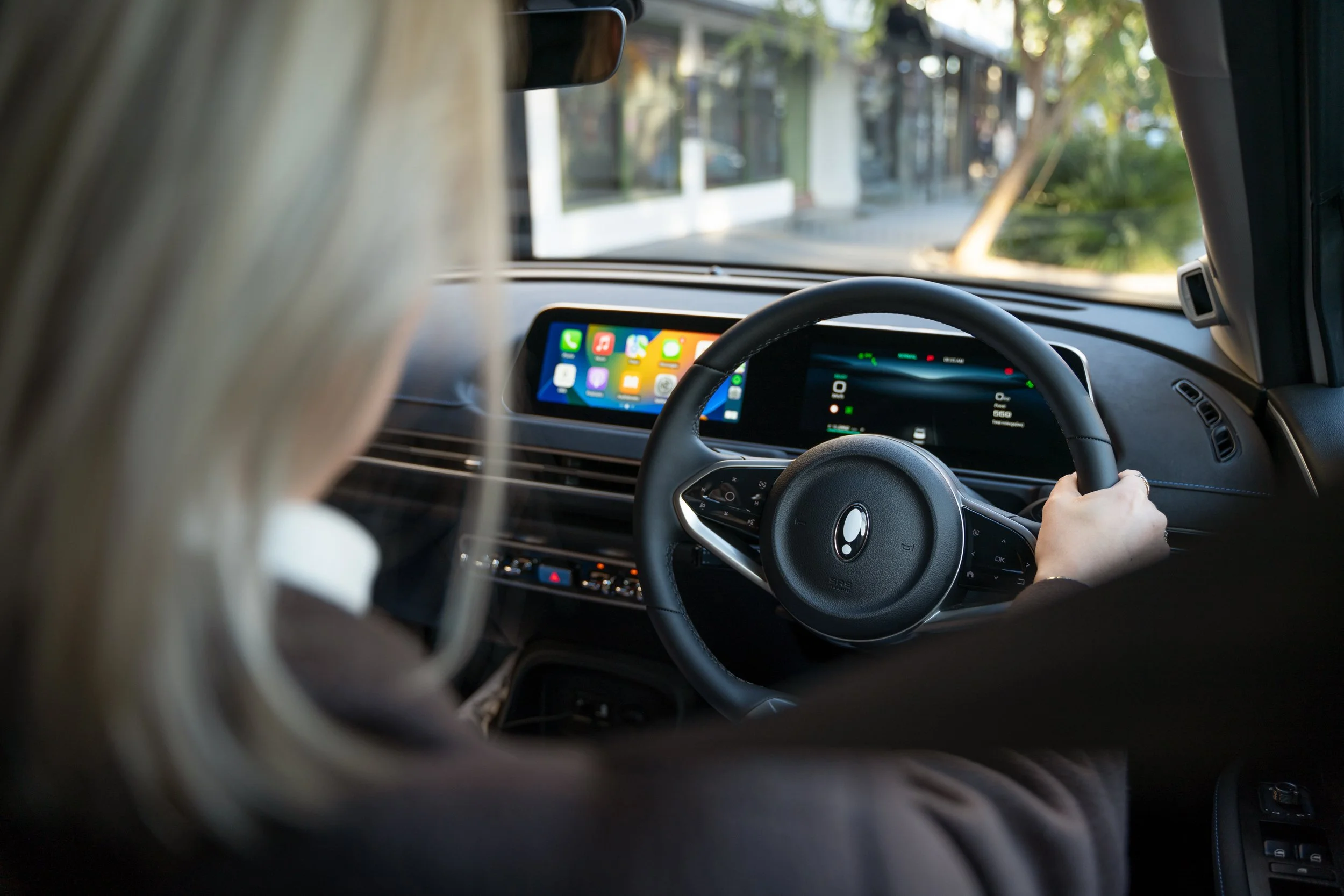 The image shows the interior of a car from the backseat perspective, with a view of the dashboard, steering wheel, and a person holding the steering wheel. A smartphone with apps displayed on the screen is mounted on the dashboard, and through the wi
