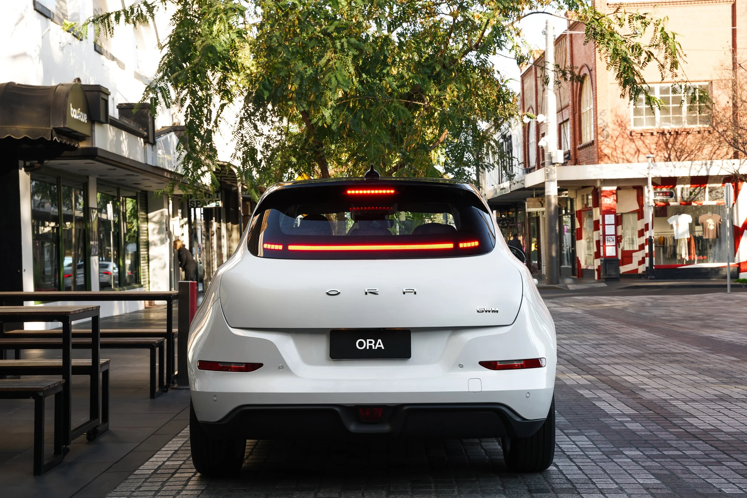 White car with the license plate 'ORA' parked on a city street in front of shops and trees, with sunlight casting shadows.