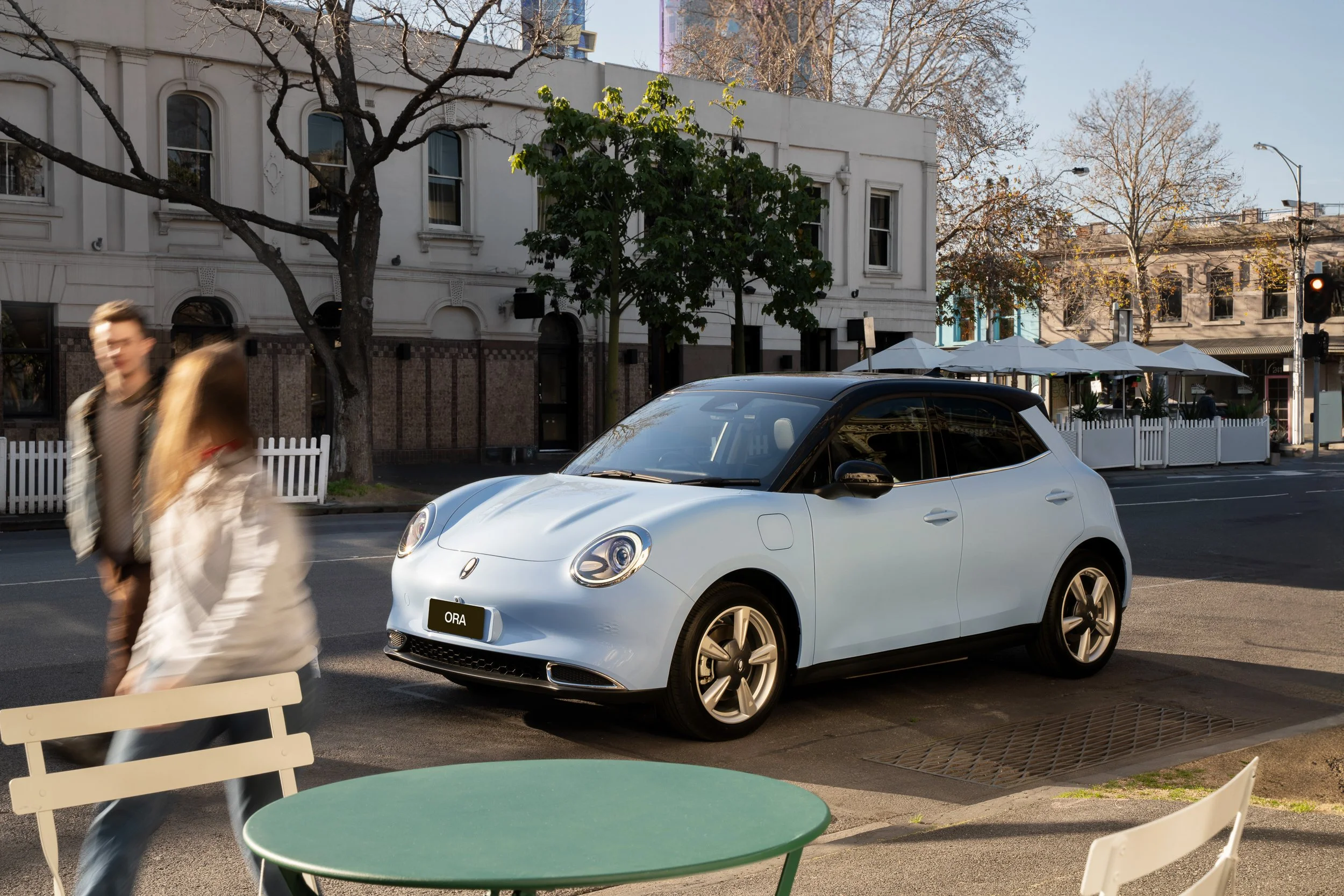 A white compact car parked on the street near sidewalk cafes with white umbrellas. Pedestrians are walking past, with some motion blur visible. Buildings and leafless trees are in the background, indicating a possible winter or autumn day.