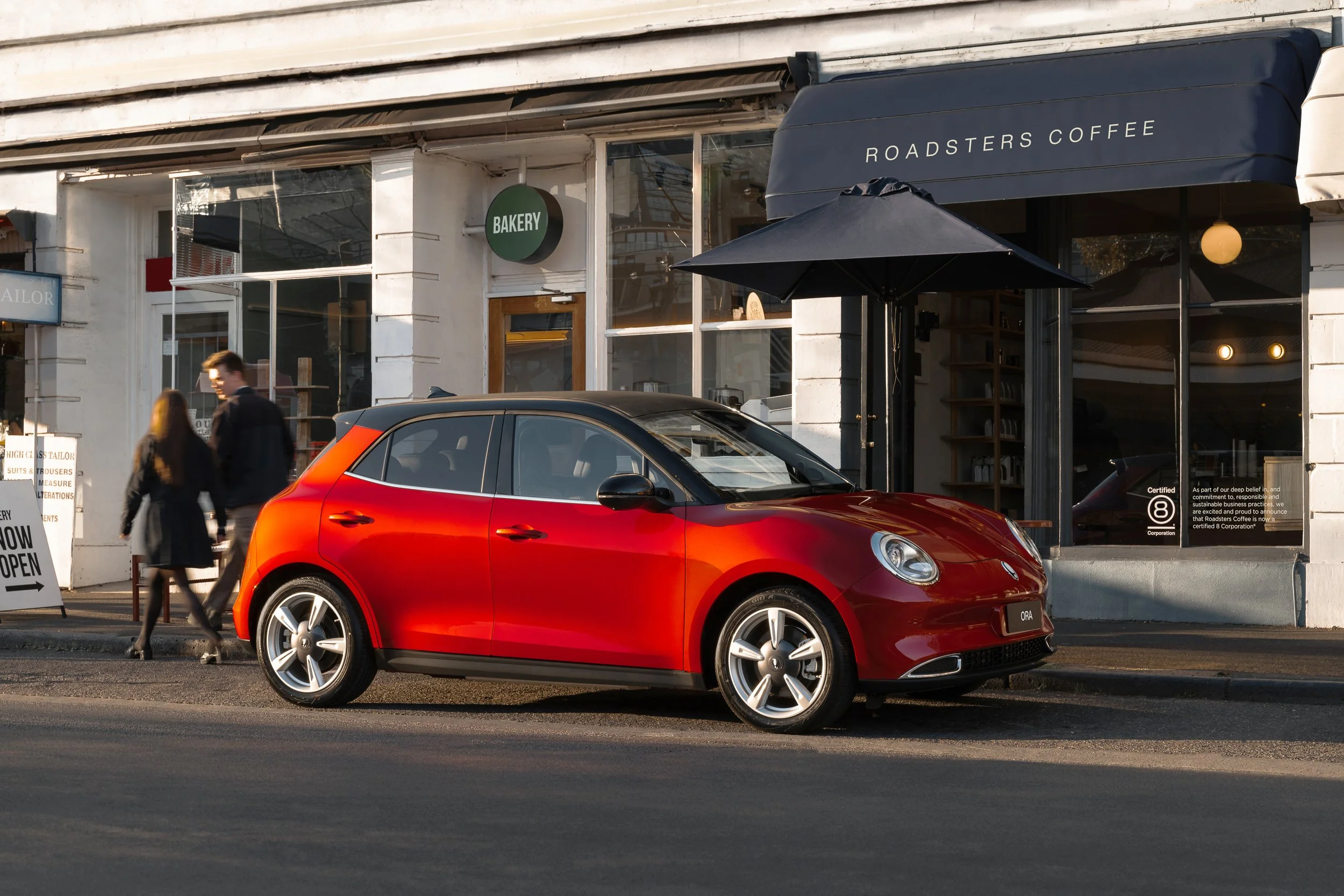 Red sports car parked on the street in front of a cafe with outdoor seating, with a person walking by and a coffee shop sign that says 'Roadsters Coffee'.