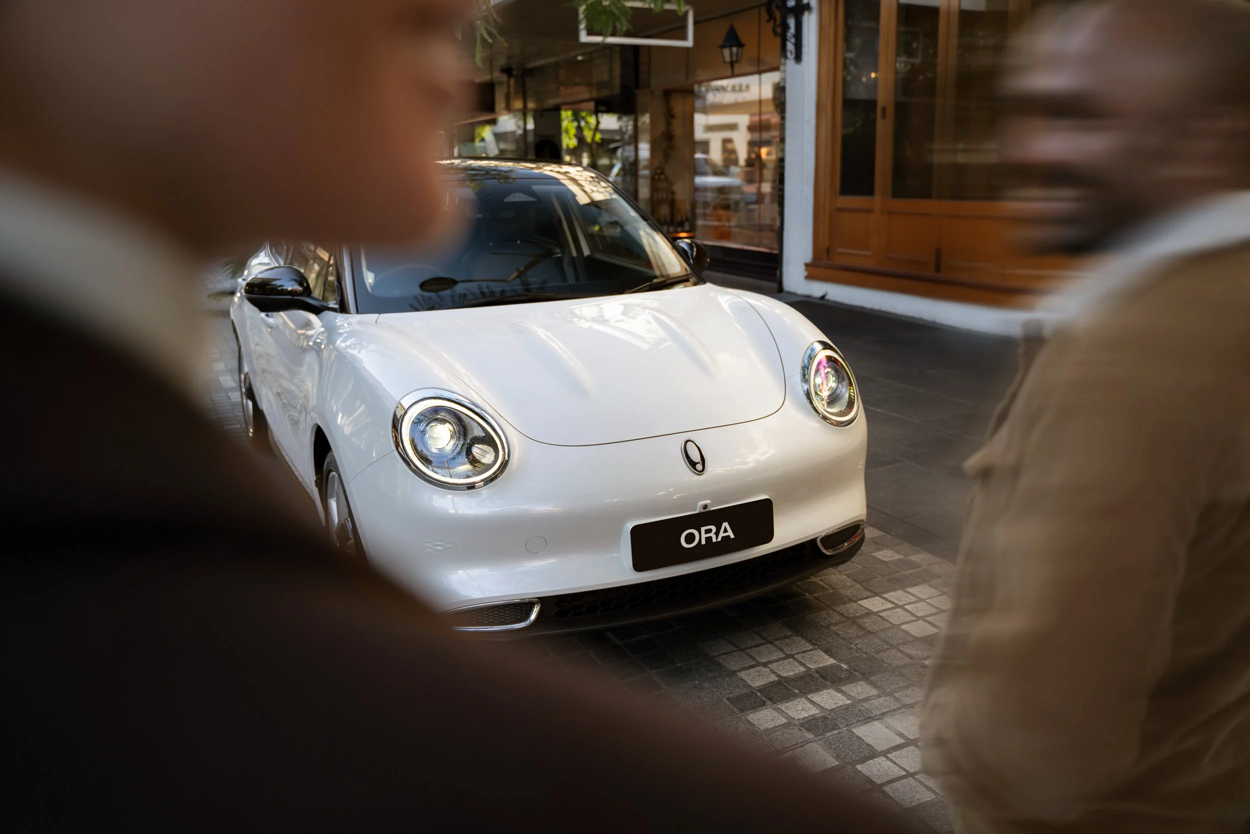 A white car parked on a city street, viewed through an opening with blurred fingers or objects in the foreground. The car has a license plate that reads 'ORA' and is reflected with sunlight. In the background, storefronts and windows are visible.