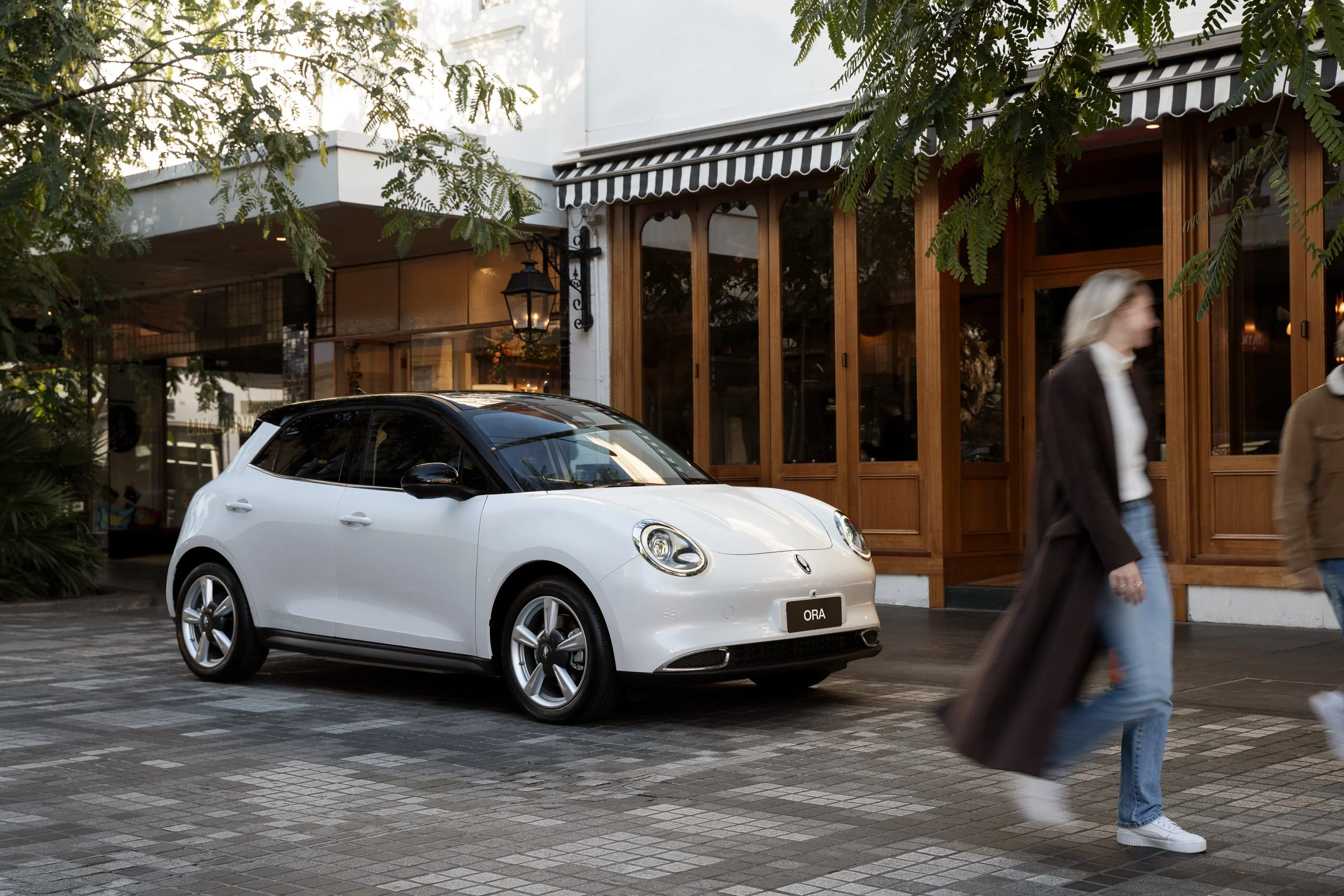 White compact car parked on a city street, with a woman walking past in front of a building with wooden framing and large windows, trees overhead, and a black and white striped awning.