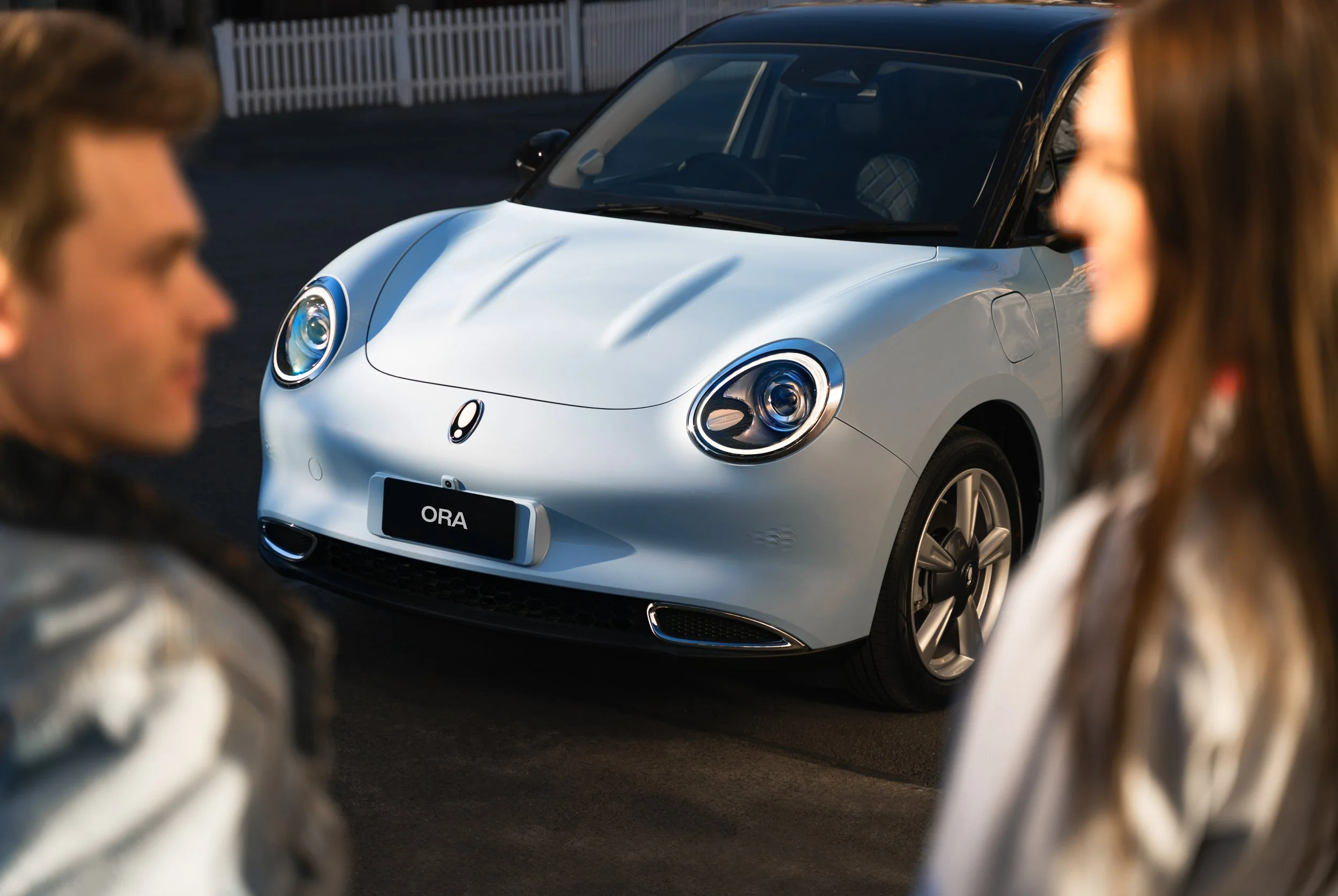 A person with brown hair and a tattoo sleeve on their arm is in the foreground on the left, and a person with long brown hair is in the foreground on the right, facing towards each other. In the background, there is a white electric vehicle with a bl