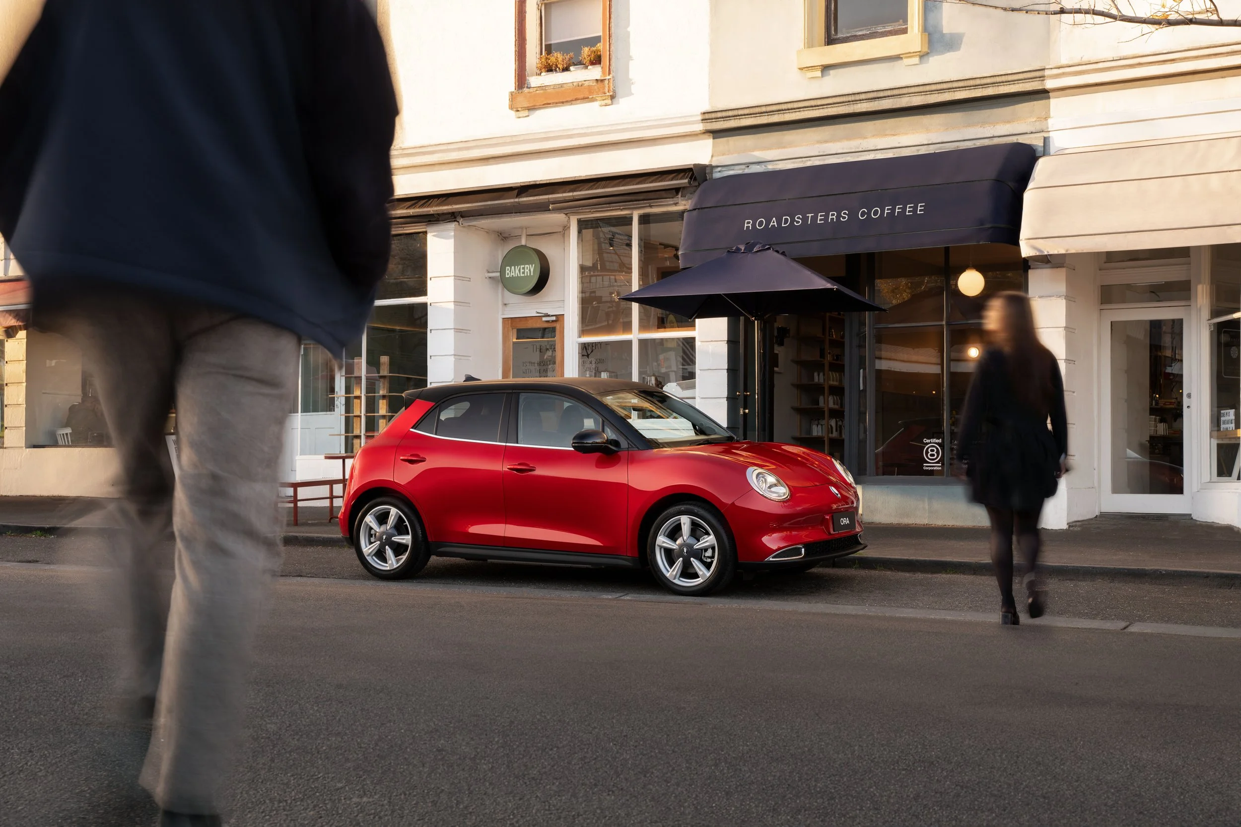 A street scene with a red sports car parked in front of a coffee shop called 'Roadsters Coffee.' There are two blurred pedestrians walking past, one on the left and one on the right, with buildings and windows in the background.