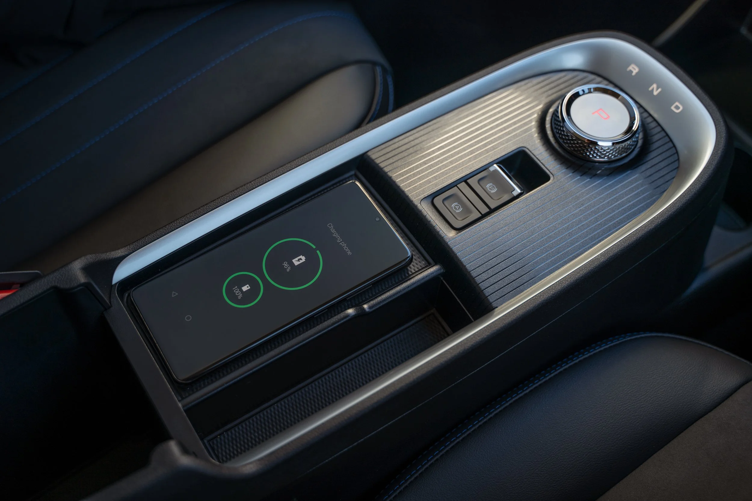 Close-up of a car's center console featuring a gear selector dial, a smartphone displaying a charging status, and other controls.