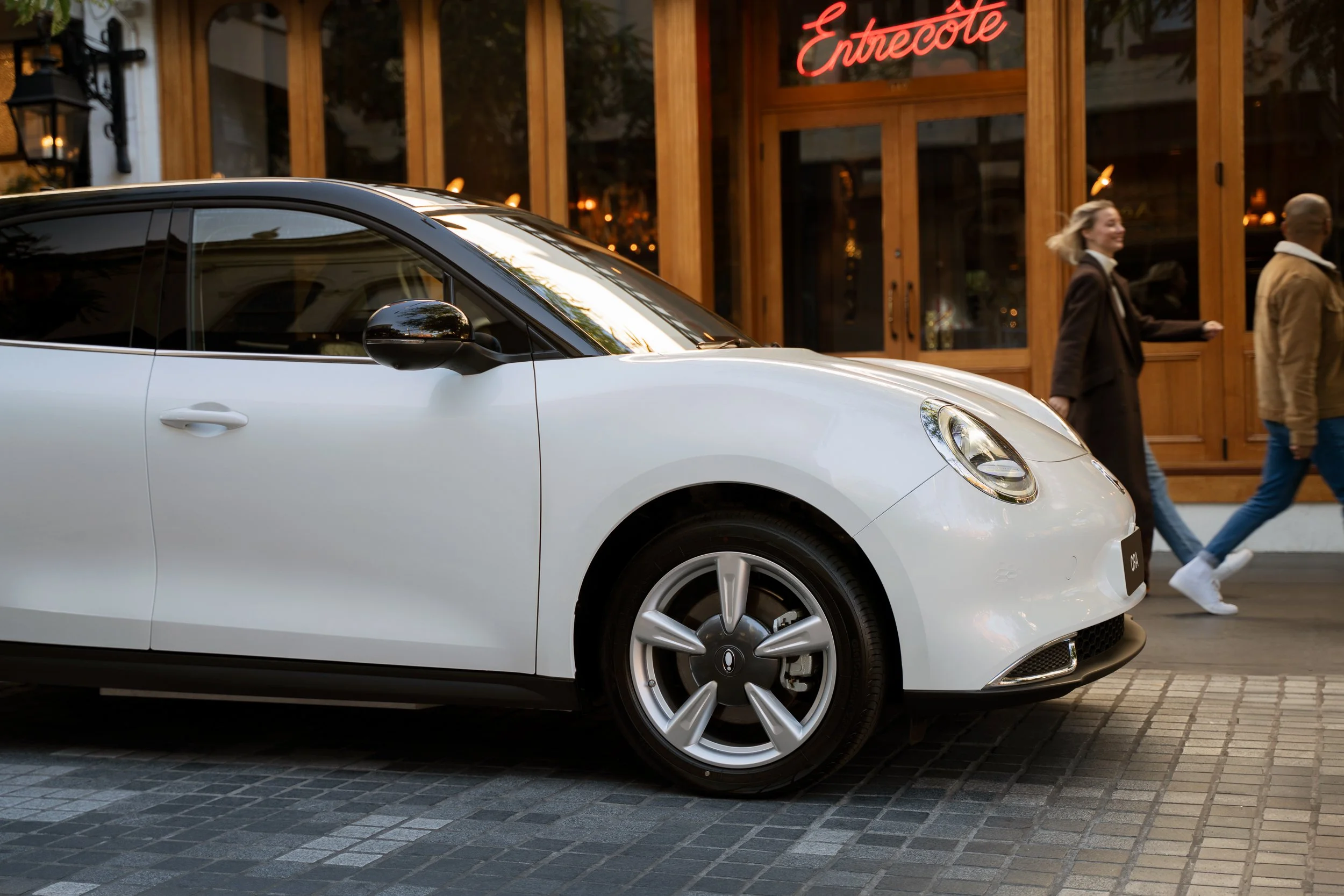 A white compact car parked on a cobblestone street in front of a wooden storefront with a red neon sign reading 'Entrecôte'. Two people are walking past the car, one woman with blonde hair and the other with a shaved head, both smiling.